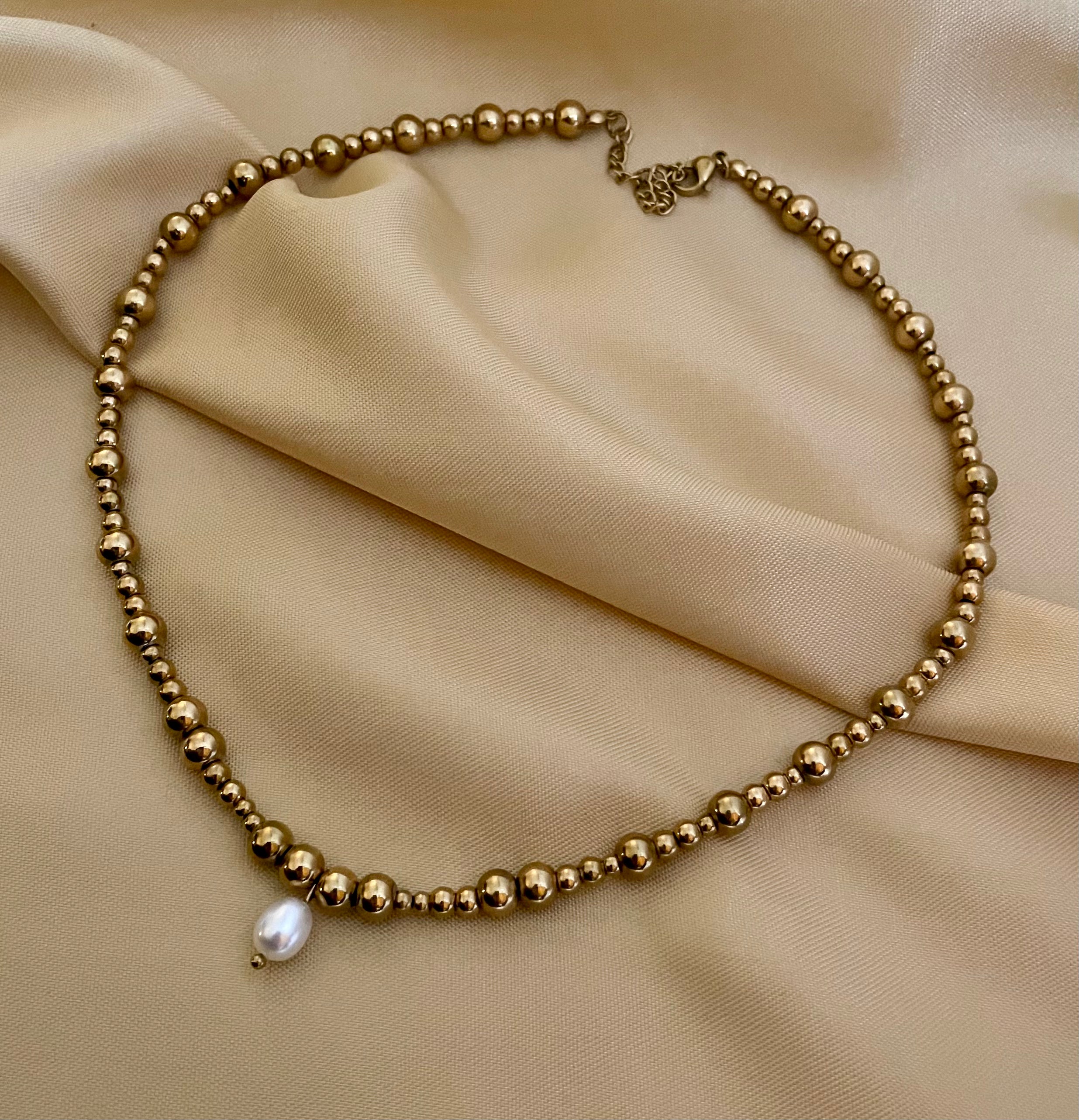 Close-up of a woman wearing a gold beaded choker necklace featuring a single white AAA grade oval pearl pendant, styled with a beige open silk shirt