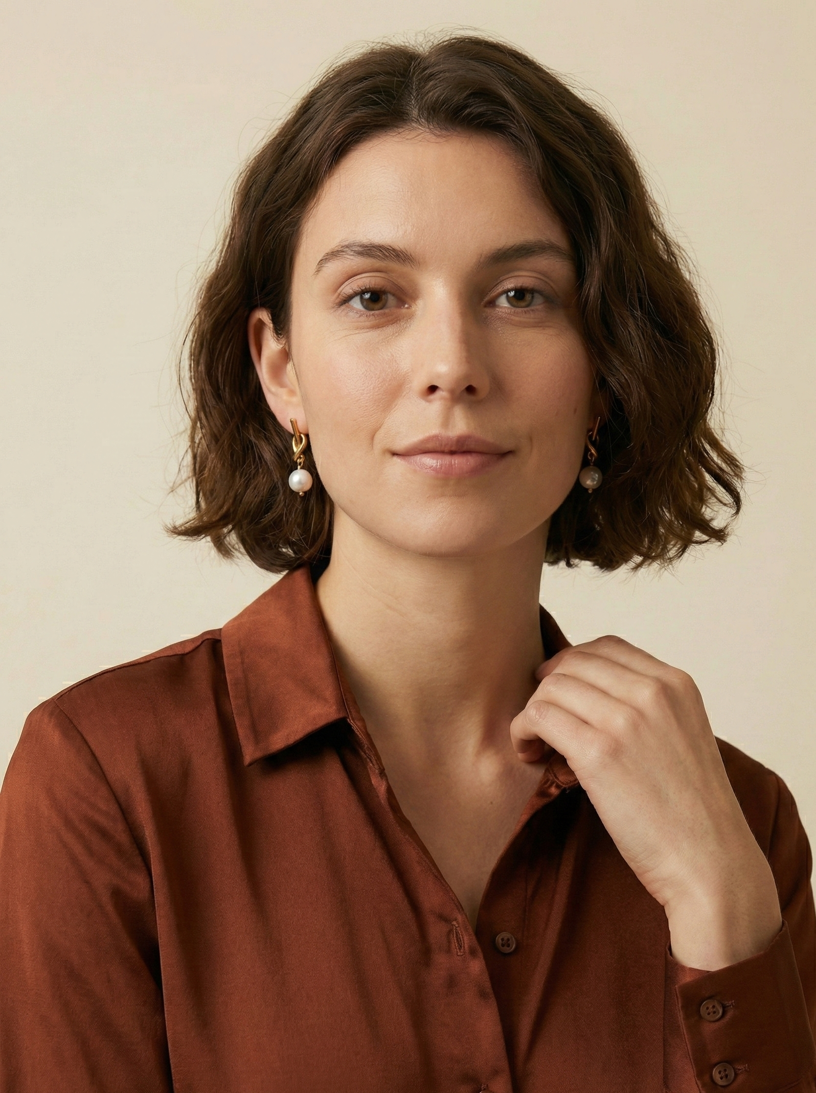 Close-up of a model wearing gold drop earrings featuring a unique knot design and a dangling white round pearl. The earrings are styled with a rust-colored silk shirt.