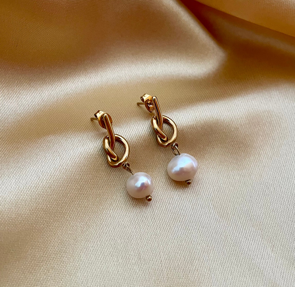 Close-up of gold drop earrings featuring a unique knot design and a dangling white round pearl.