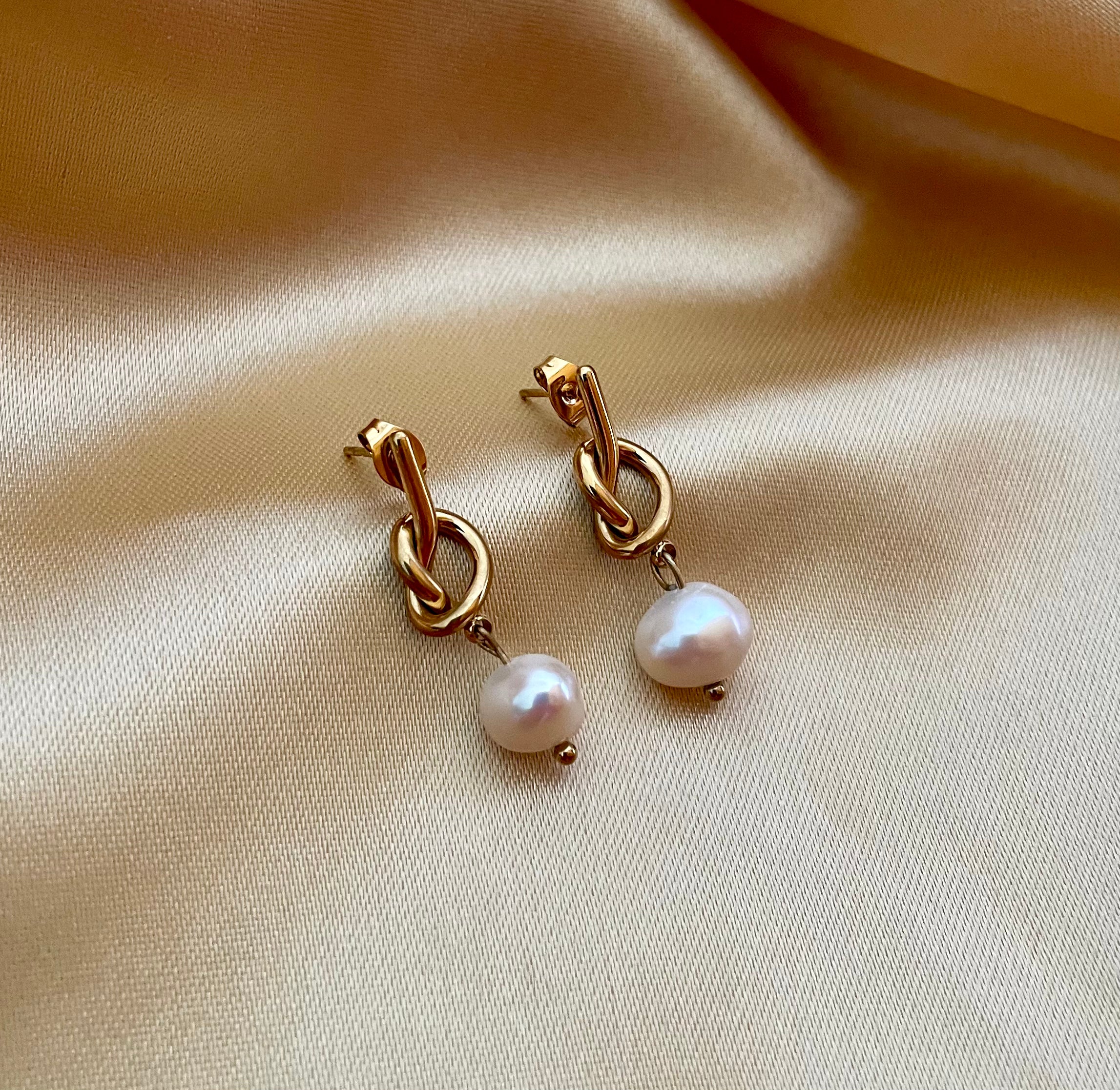 Close-up of gold drop earrings featuring a unique knot design and a dangling white round pearl.