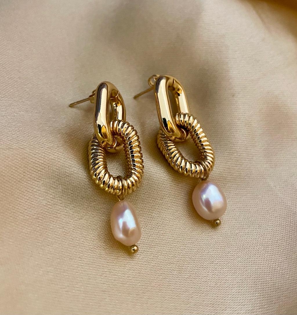 Close-up of sculptural gold link earrings featuring a twisted rope texture and hanging freshwater baroque pearl drops. The earrings are displayed on a champagne silk fabric to highlight the pearl luster and high-shine gold finish
