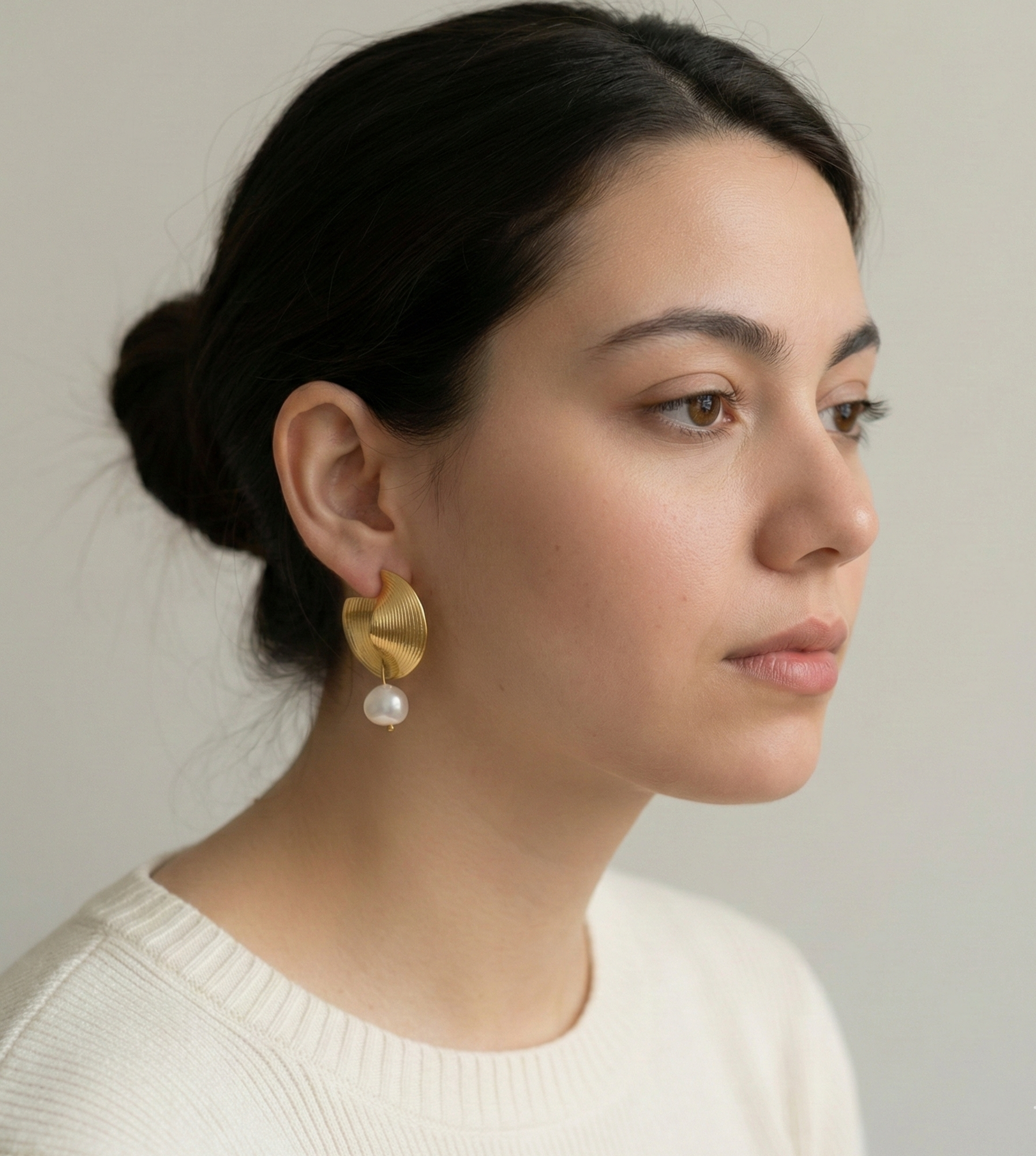 Side profile of a model wearing textured gold fan-shaped earrings with a hanging freshwater pearl and natural hair.