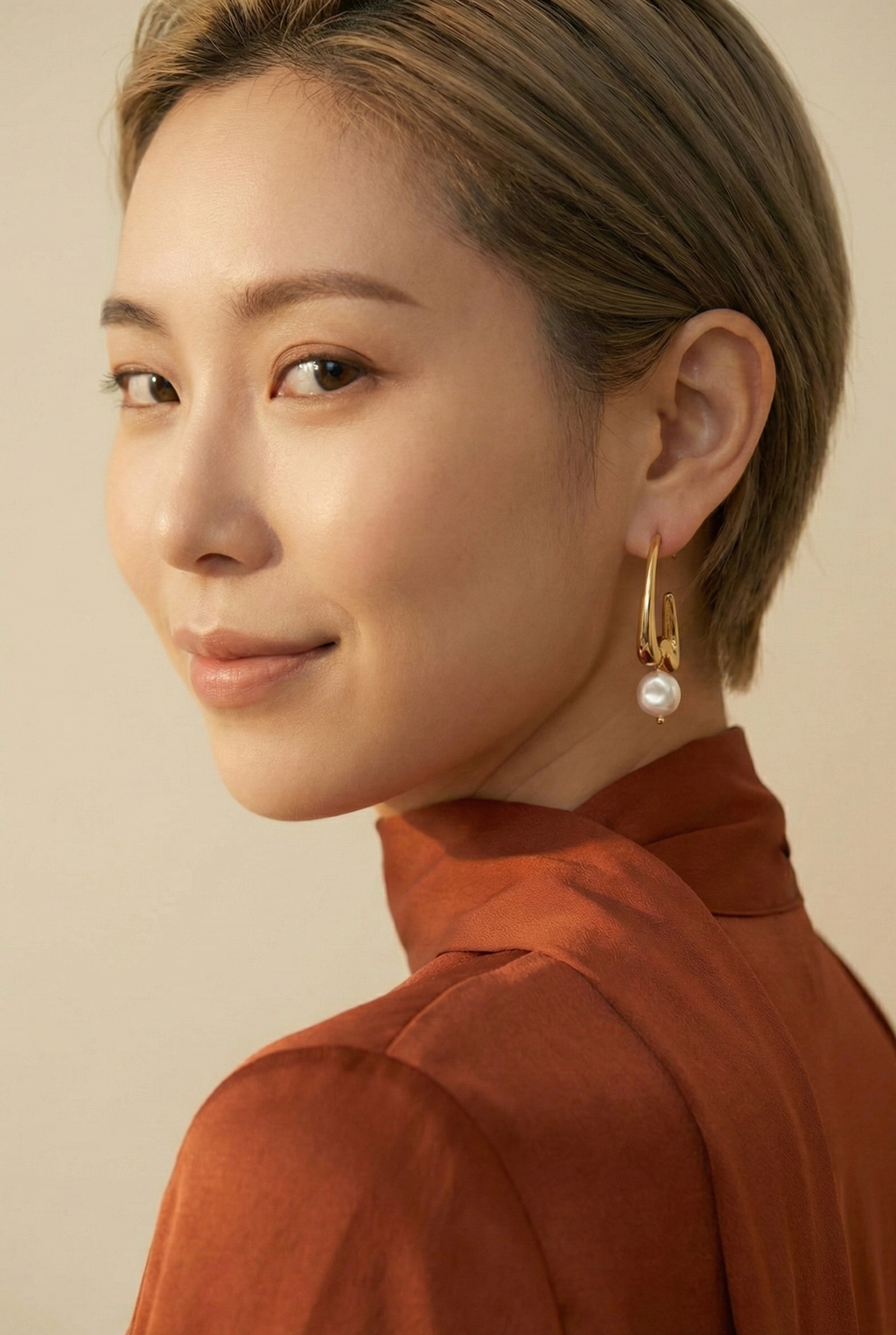 Close-up of a person wearing a gold hoop earring with a pearl, against a neutral background.