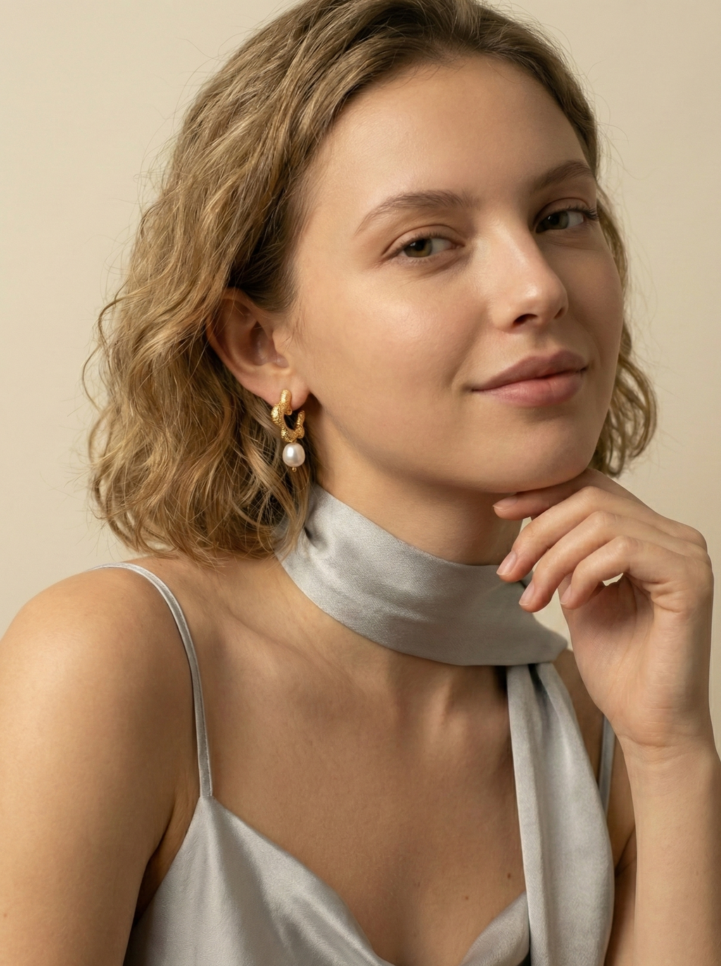 Model wearing small textured gold hoop earrings with a single round white pearl drop and a grey silk neck scarf.