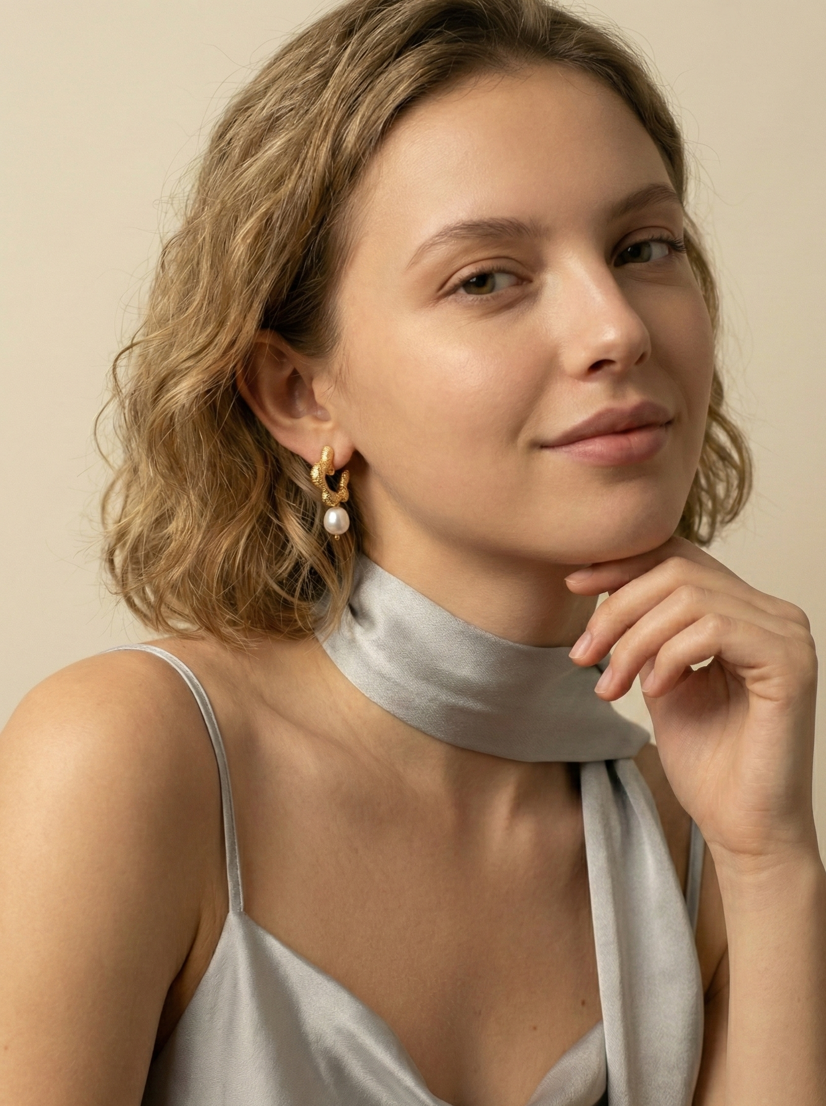 Model wearing small textured gold hoop earrings with a single round white pearl drop and a grey silk neck scarf.