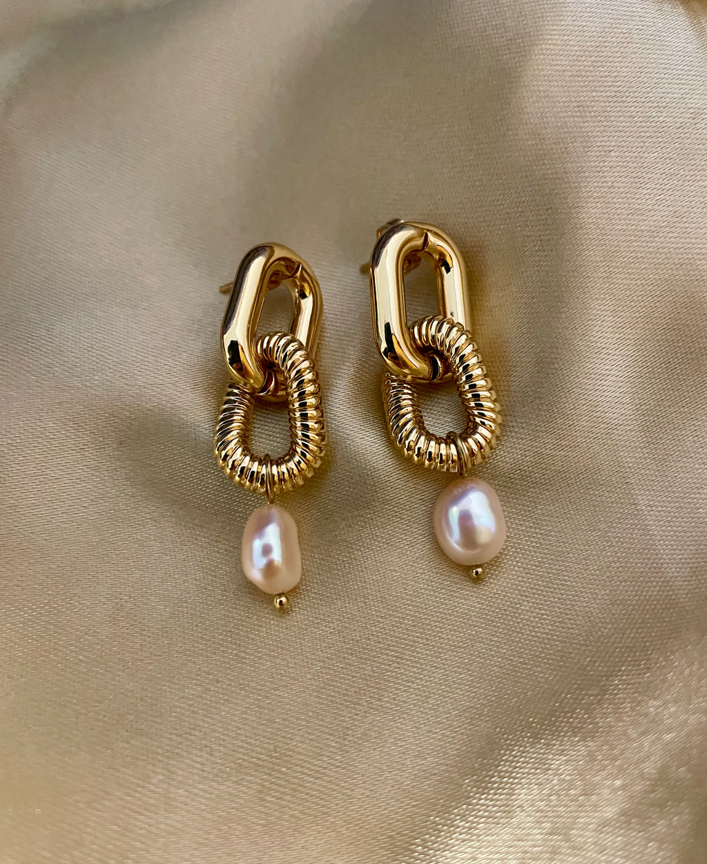 Close-up of sculptural gold link earrings featuring a twisted rope texture and hanging pink freshwater baroque pearl drops. The earrings are displayed on a champagne silk fabric to highlight the pearl luster and high-shine gold finish
