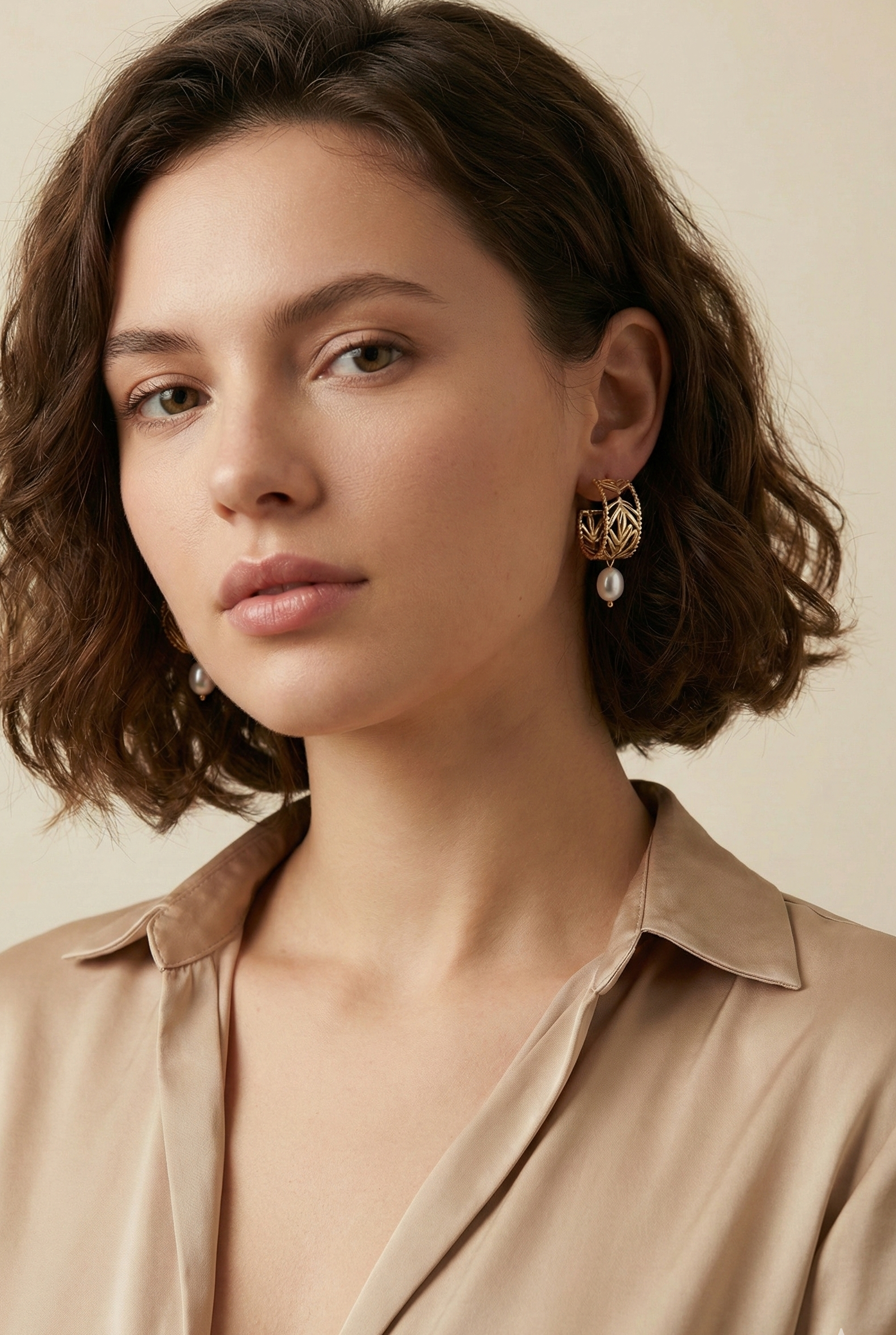 Close-up editorial profile of a model wearing detailed gold openwork hoop earrings featuring a leaf motif and a single dangling white oval pearl. The warm, soft lighting highlights the pearl's luster and the gold texture against the model's skin, set against a neutral background.