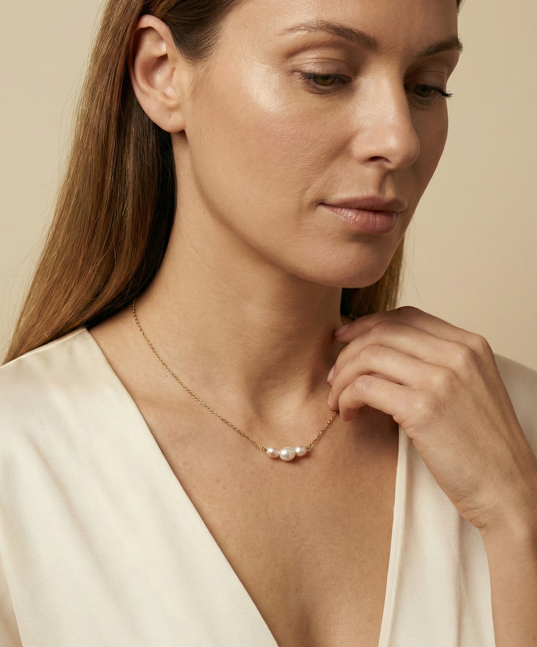 Close-up of a model wearing a dainty gold chain necklace featuring a trio of small pearls: a central white pearl flanked by two pink oval baroque pearls, styled with an ivory silk top