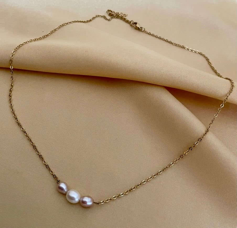 Close-up of a model wearing a dainty gold chain necklace featuring a trio of small pearls: a central white pearl flanked by two pink oval baroque pearls, styled with an ivory silk top