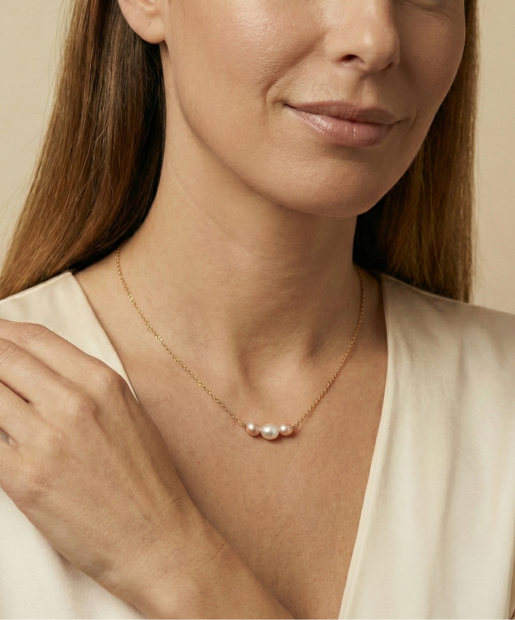 Close-up of a model wearing a dainty gold chain necklace featuring a trio of small pearls: a central white pearl flanked by two pink oval baroque pearls, styled with an ivory silk top