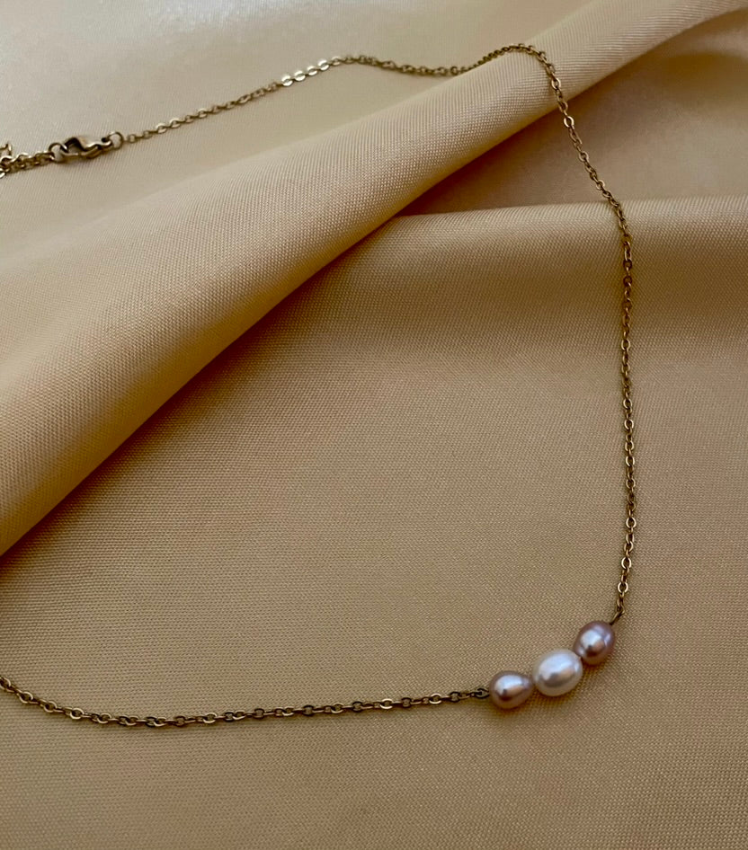 Close-up of a model wearing a dainty gold chain necklace featuring a trio of small pearls: a central white pearl flanked by two pink oval baroque pearls, styled with an ivory silk top