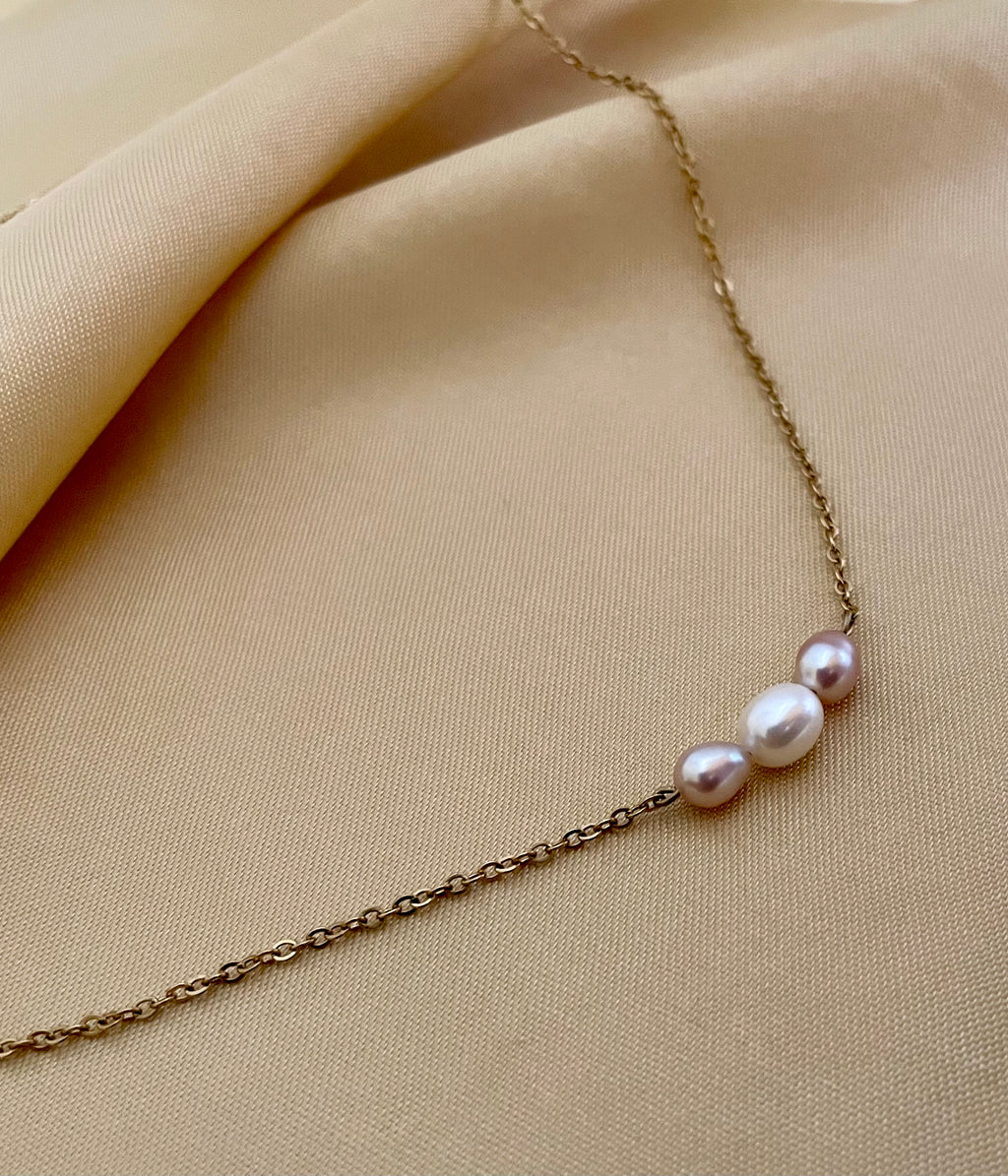 Close-up of a model wearing a dainty gold chain necklace featuring a trio of small pearls: a central white pearl flanked by two pink oval baroque pearls, styled with an ivory silk top