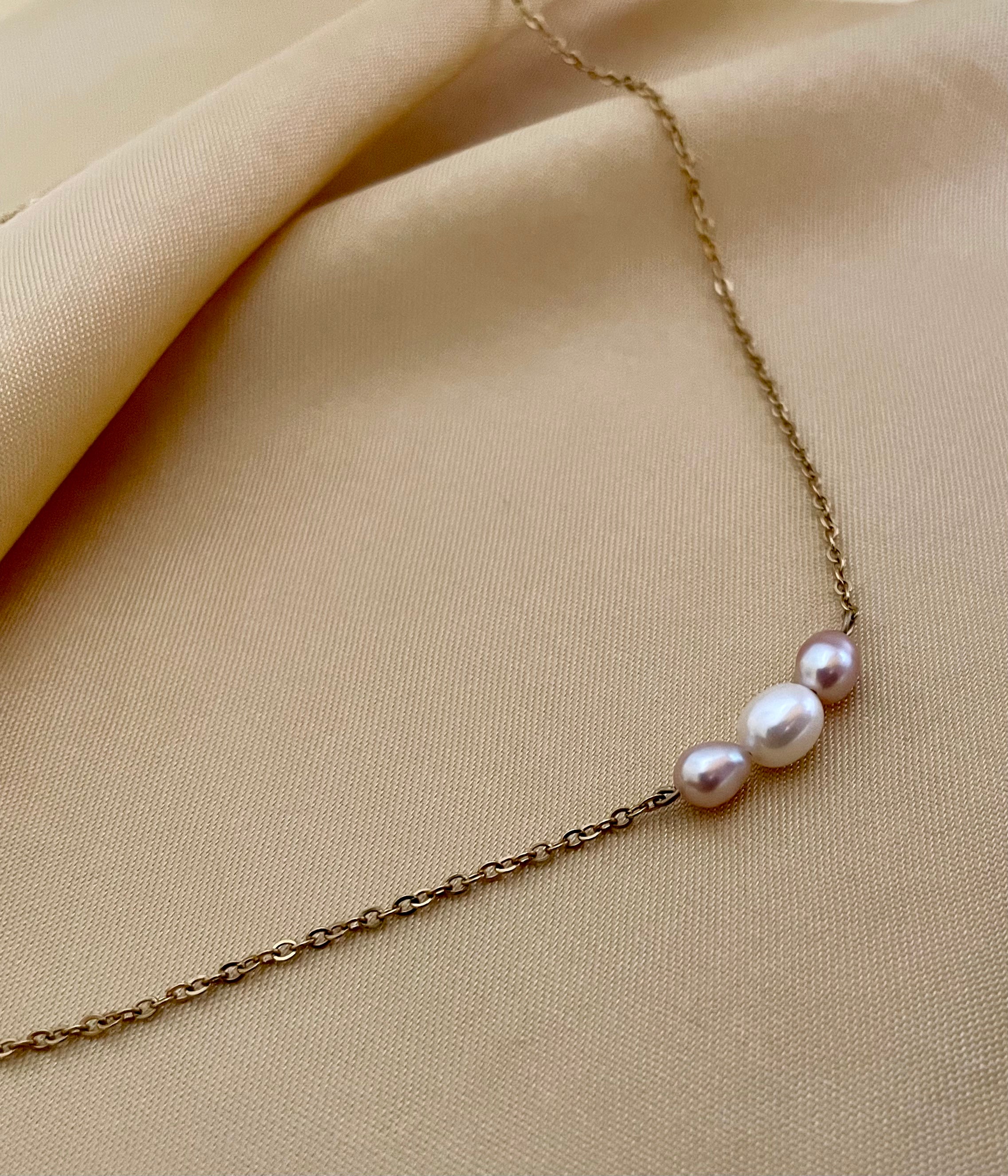 Close-up of a model wearing a dainty gold chain necklace featuring a trio of small pearls: a central white pearl flanked by two pink oval baroque pearls, styled with an ivory silk top