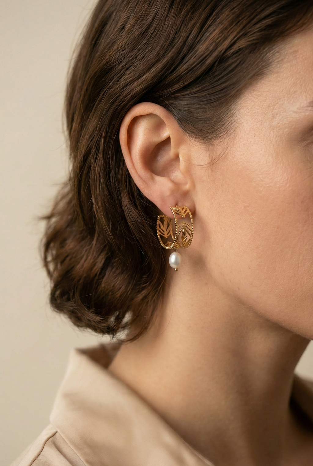 Close-up editorial profile of a model wearing detailed gold openwork hoop earrings featuring a leaf motif and a single dangling white oval pearl. The warm, soft lighting highlights the pearl's luster and the gold texture against the model's skin, set against a neutral background.