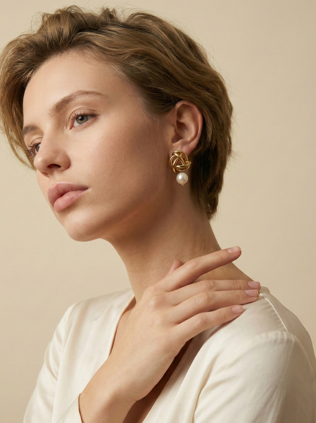 Close-up of gold twisted knot earrings with a white pearl drop, worn by a model in a cream silk blouse