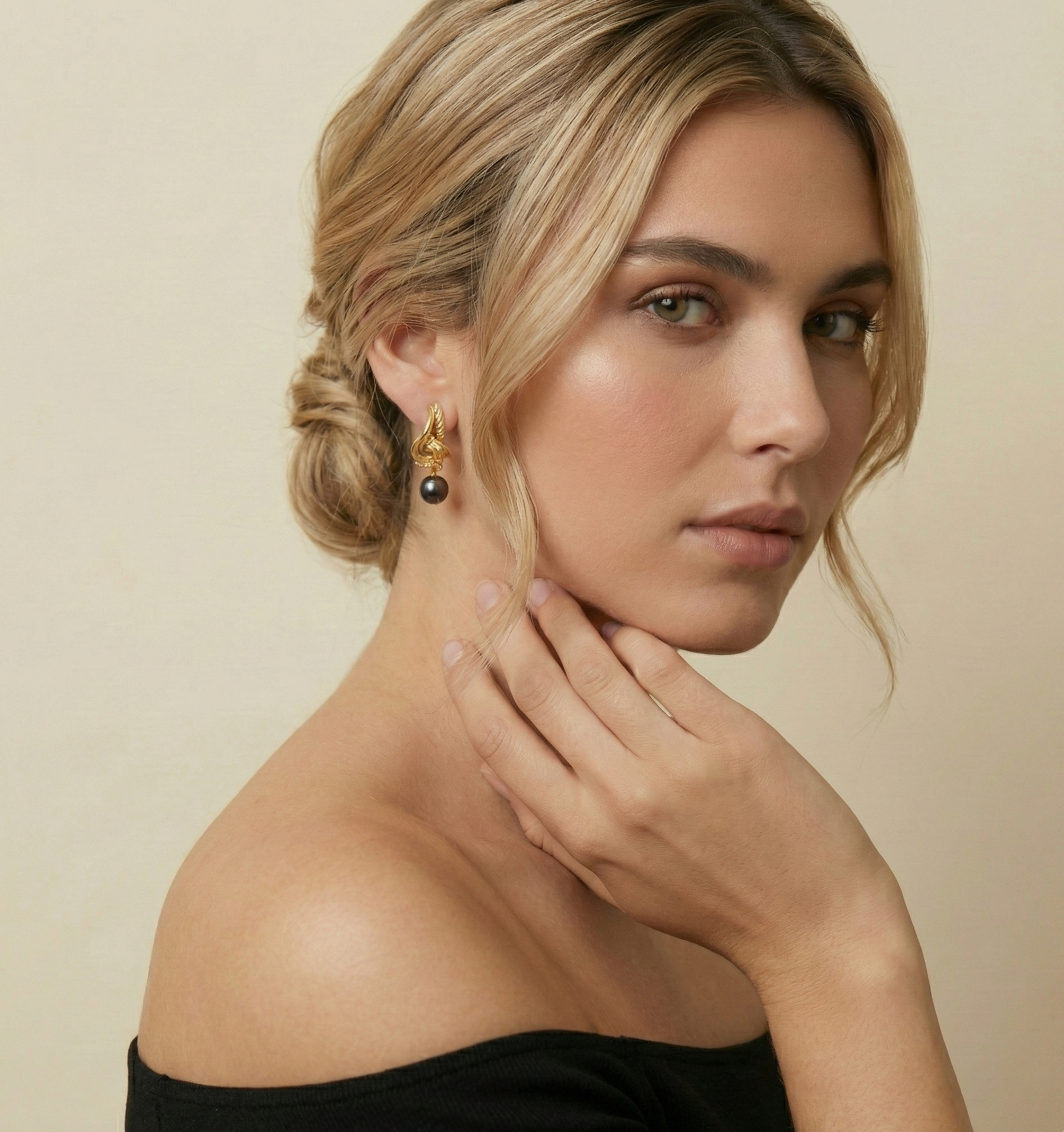 Luxury gold knot hoop earrings with round black pearl drop. High-fashion jewelry photography showing elegant styling with an off-shoulder black top and classic hairstyle. Perfect for evening wear and sophisticated looks