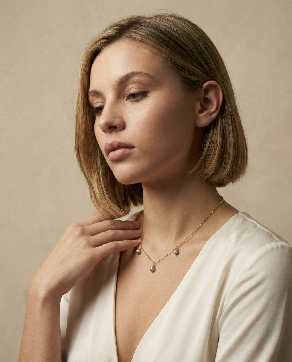 Close-up detail of a delicate gold chain necklace featuring three small metallic grey baroque pearls with high luster, draped elegantly on a woman's collarbone against an ivory silk top. High-resolution jewelry photography highlighting the pearl texture and dainty chain links