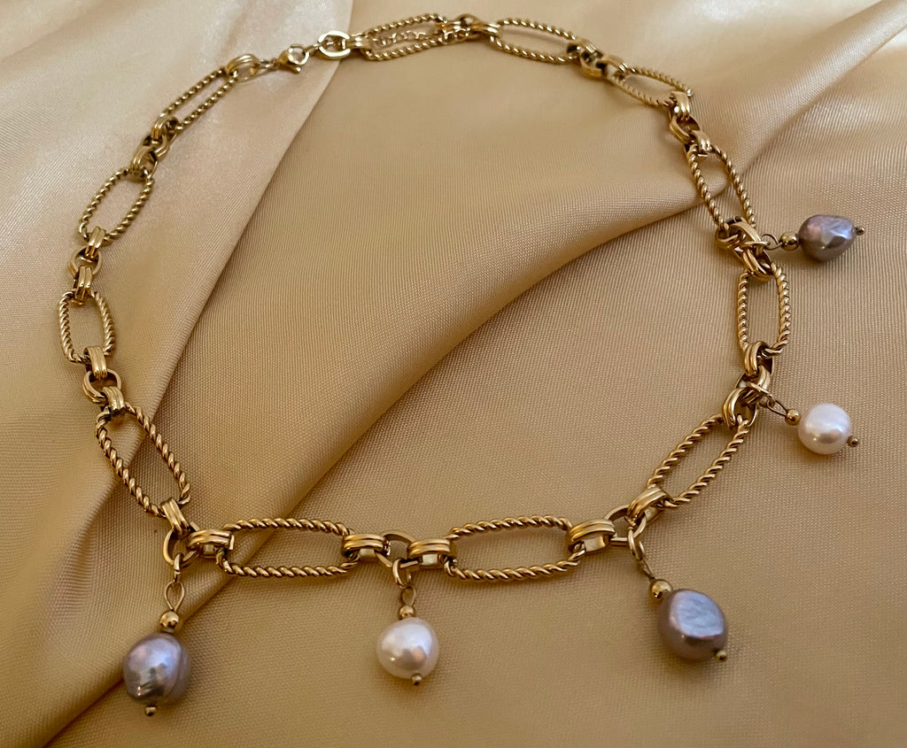 Close-up of a gold link chain necklace featuring dangling white and grey baroque pearl charms on a model wearing an ivory coa