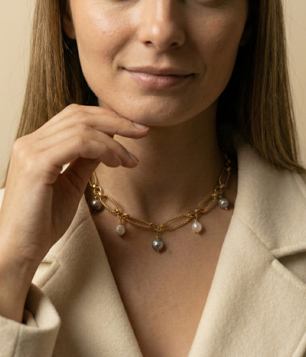 Close-up of a gold link chain necklace featuring dangling white and grey baroque pearl charms on a model wearing an ivory coa