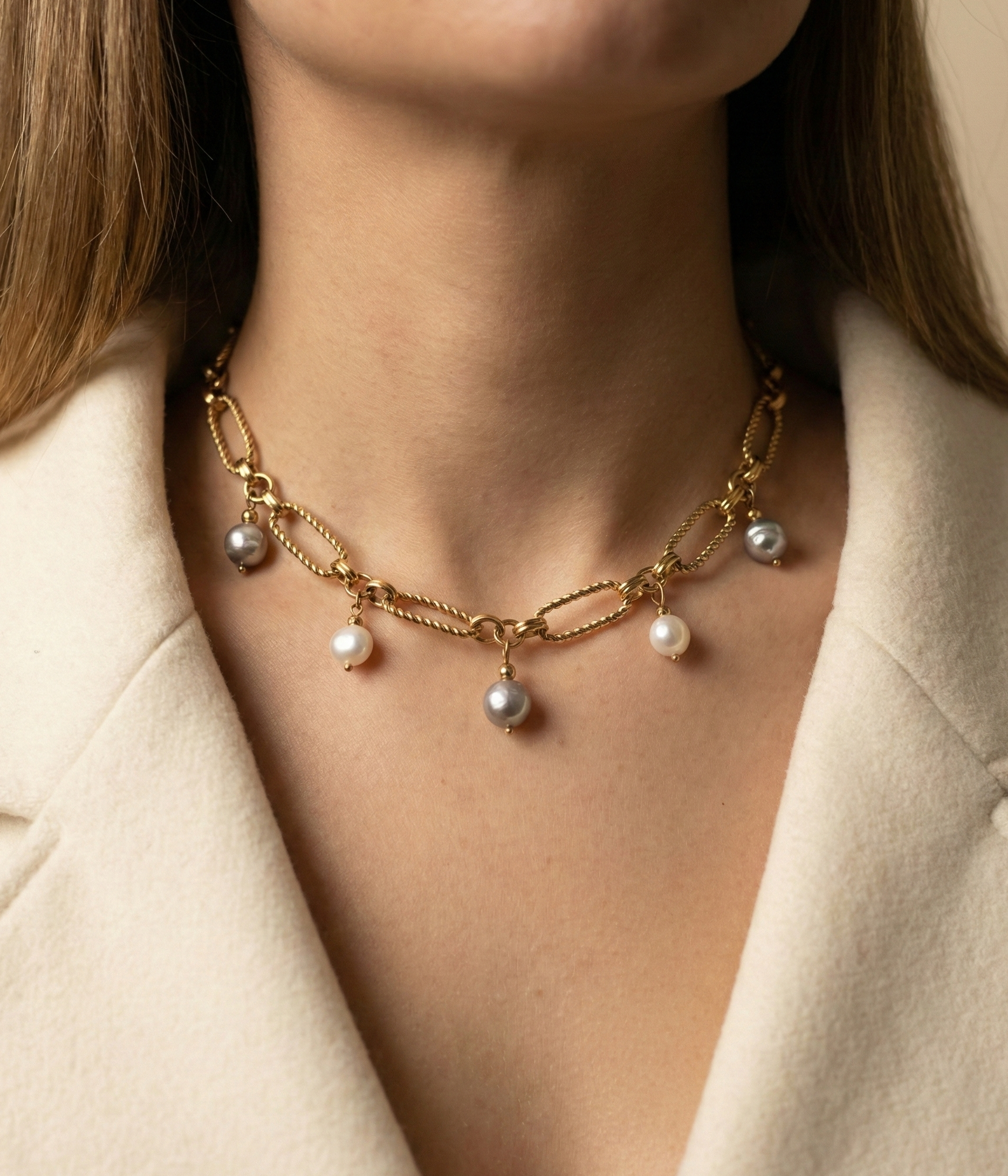 Close-up of a gold link chain necklace featuring dangling white and grey baroque pearl charms on a model wearing an ivory coa