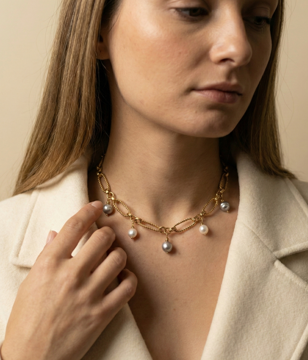 Close-up of a gold link chain necklace featuring dangling white and grey baroque pearl charms on a model wearing an ivory coa