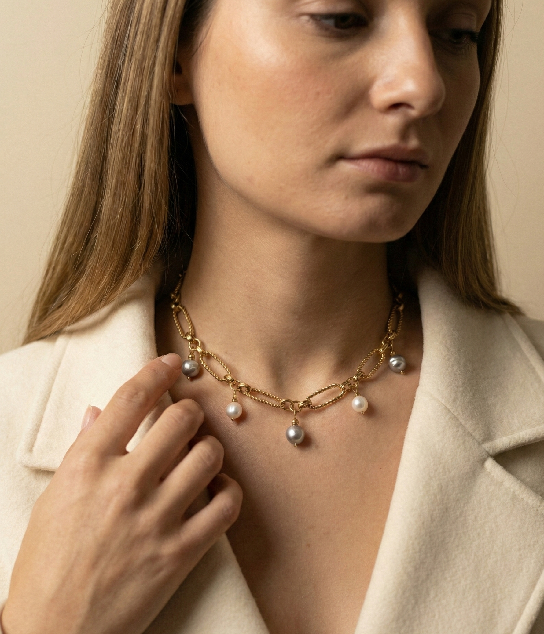 Close-up of a gold link chain necklace featuring dangling white and grey baroque pearl charms on a model wearing an ivory coa