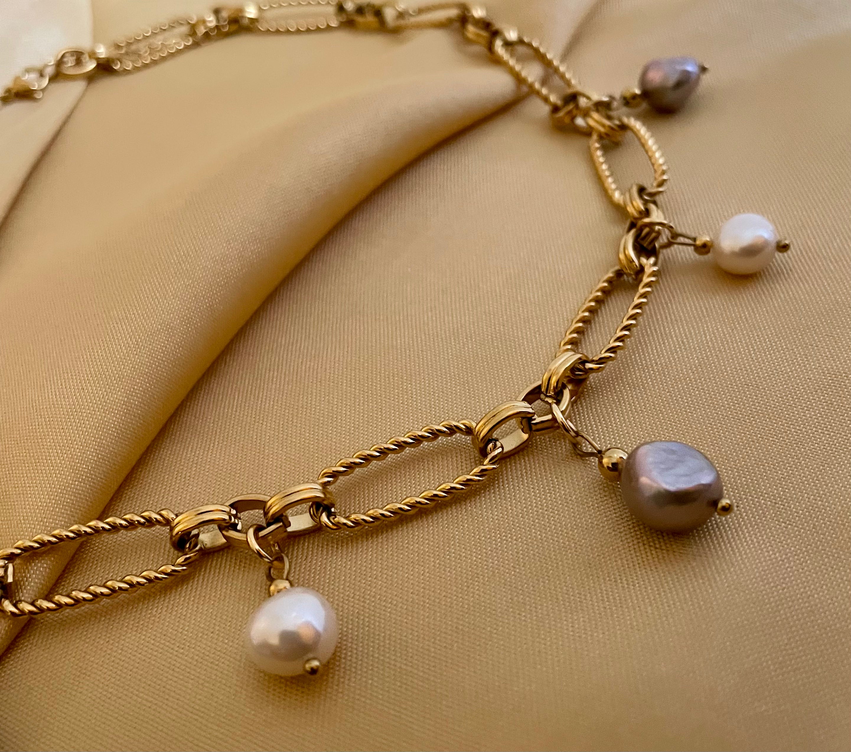 Close-up of a gold link chain necklace featuring dangling white and grey baroque pearl charms on a model wearing an ivory coa
