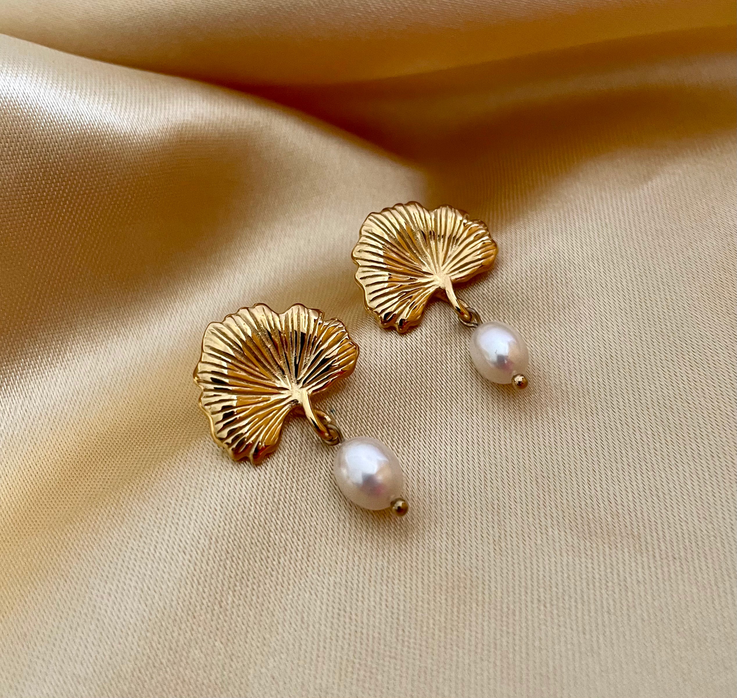 The earrings feature a detailed, textured gold fan shape resembling a ginkgo biloba leaf, from which hangs a lustrous white freshwater pearl.