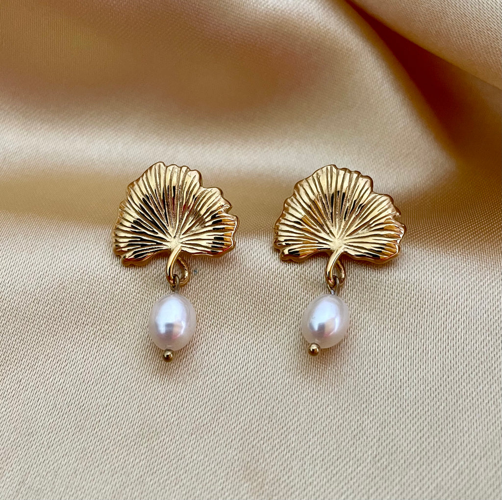 The earrings feature a detailed, textured gold fan shape resembling a ginkgo biloba leaf, from which hangs a lustrous white freshwater pearl.