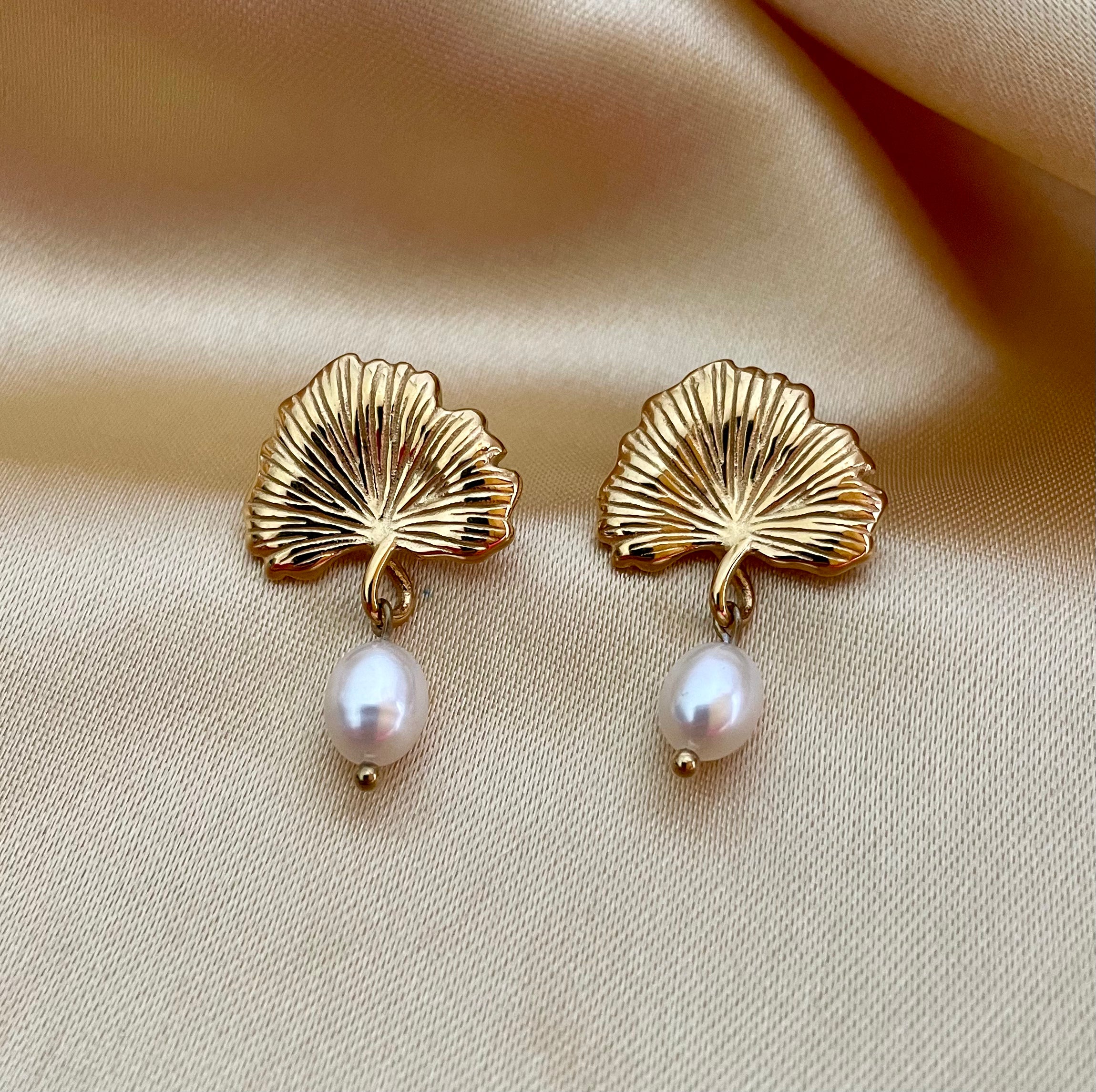 The earrings feature a detailed, textured gold fan shape resembling a ginkgo biloba leaf, from which hangs a lustrous white freshwater pearl.