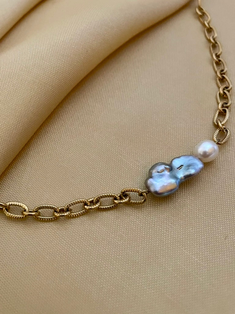 Close-up editorial portrait of a woman wearing a gold oval-link chain necklace featuring a grey baroque keshi pearl and a small white pearl, styled with a deep V-neck ivory silk top