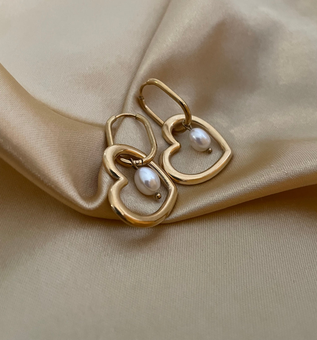 Close-up of a model wearing gold heart-shaped hoop earrings with a lustrous white pearl charm. High-quality jewelry photography showcasing the polished gold finish and pearl texture against a plain ivory background