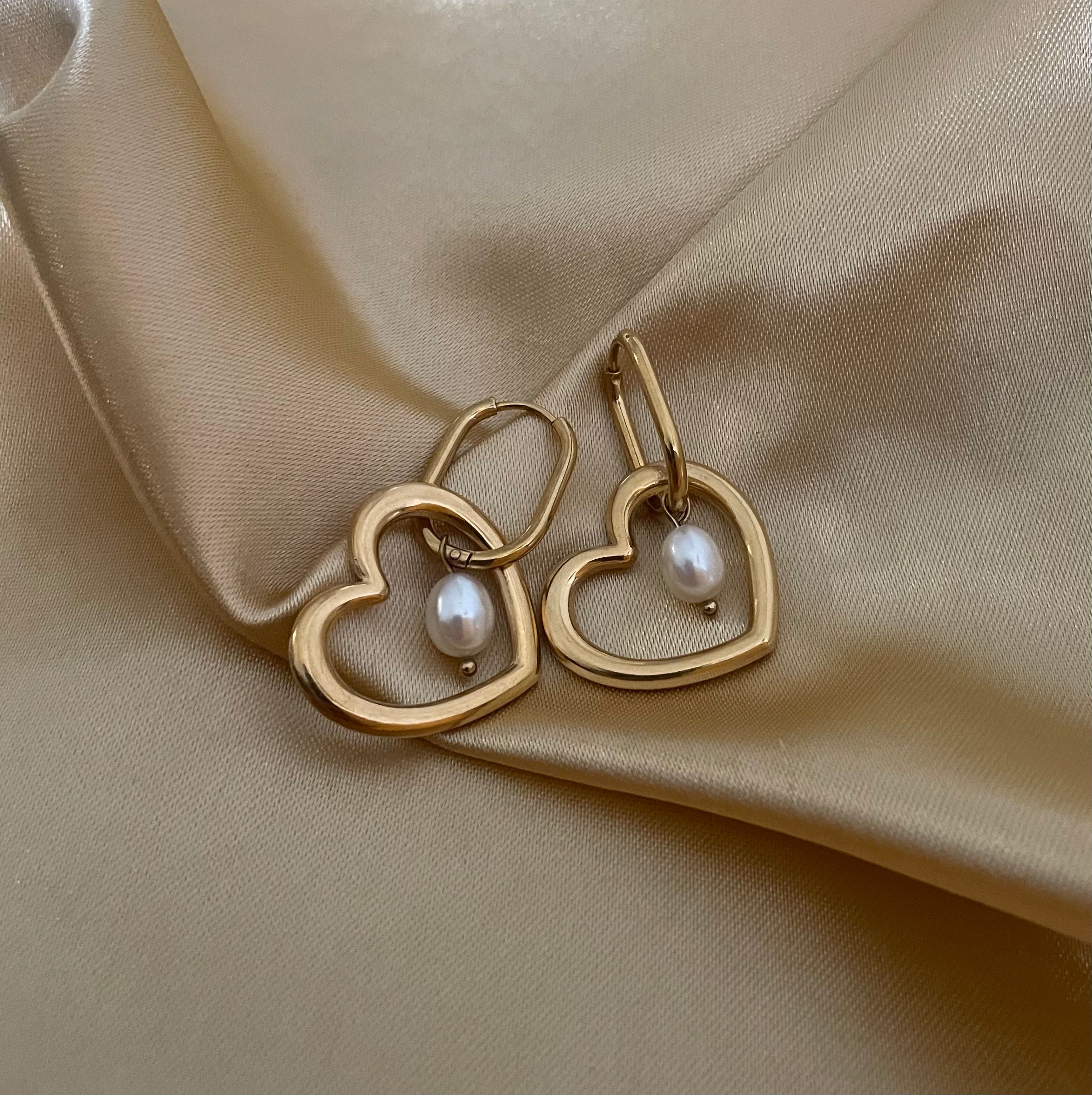 Close-up of a model wearing gold heart-shaped hoop earrings with a lustrous freshwater white pearl charm. High-quality jewelry photography showcasing the polished gold finish and pearl texture against a plain ivory background