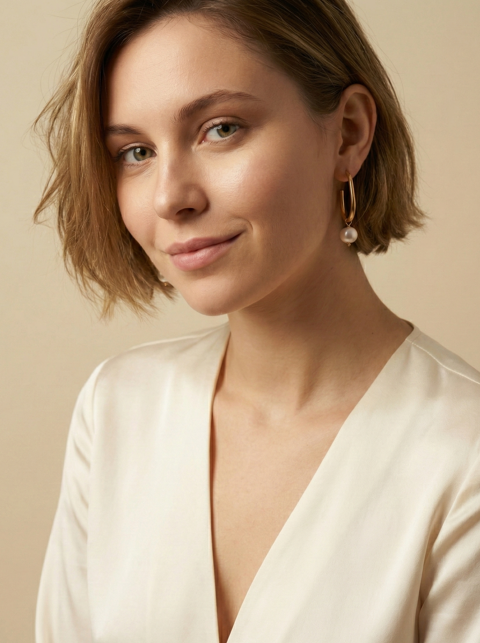 Close-up side profile of a woman's ear wearing a gold hoop earring with a single white pearl drop, highlighting the metallic luster and pearl detail against a soft neutral background