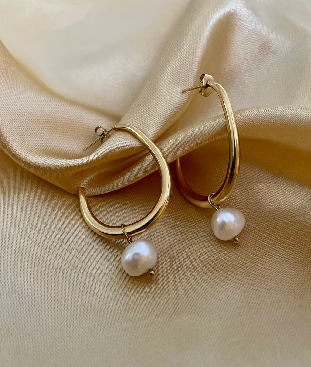 Close-up of a gold hoop earring with a single white pearl drop, highlighting the metallic luster and pearl detail against a soft neutral background