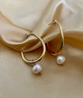 Close-up of a gold hoop earring with a single white pearl drop, highlighting the metallic luster and pearl detail against a soft neutral background