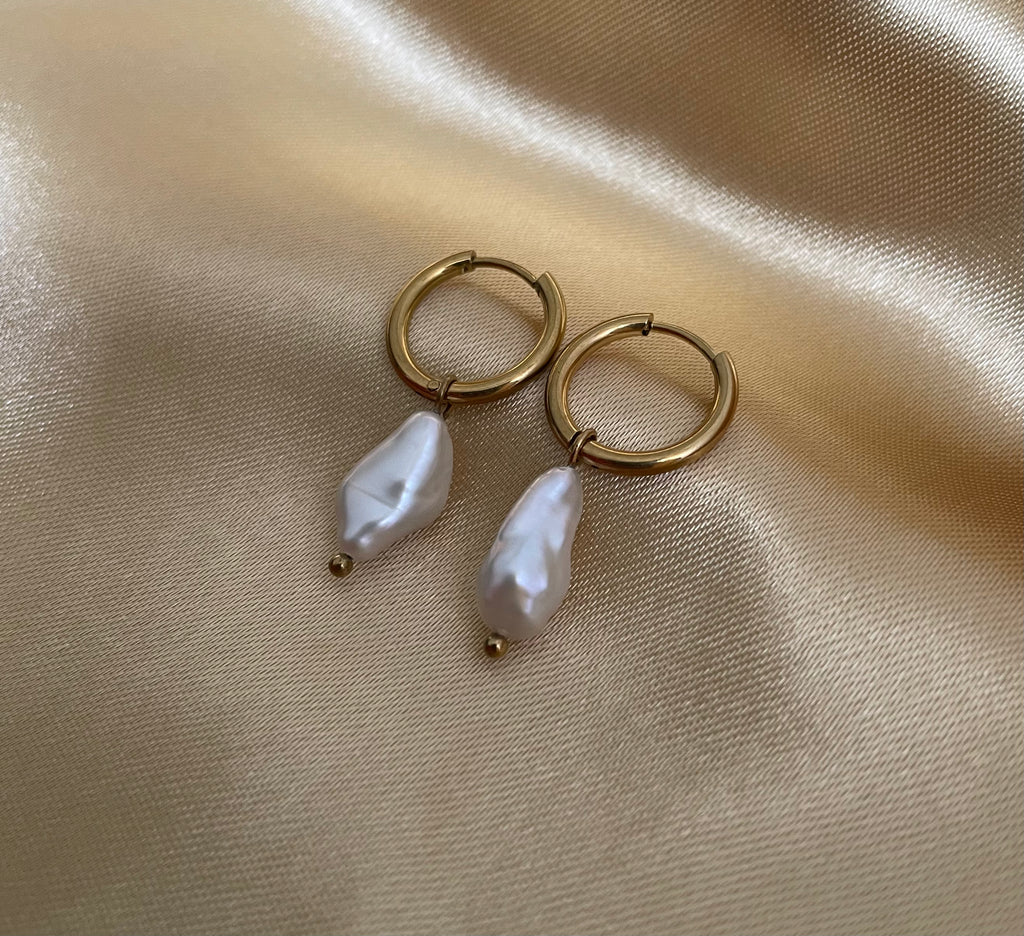 Close-up photography of a dainty gold hoop earring featuring a lustrous white baroque pearl drop. High-resolution commercial jewelry shot showcasing organic keshi pearl texture, nacre iridescence, and a simple gold clasp against a matte ivory background.