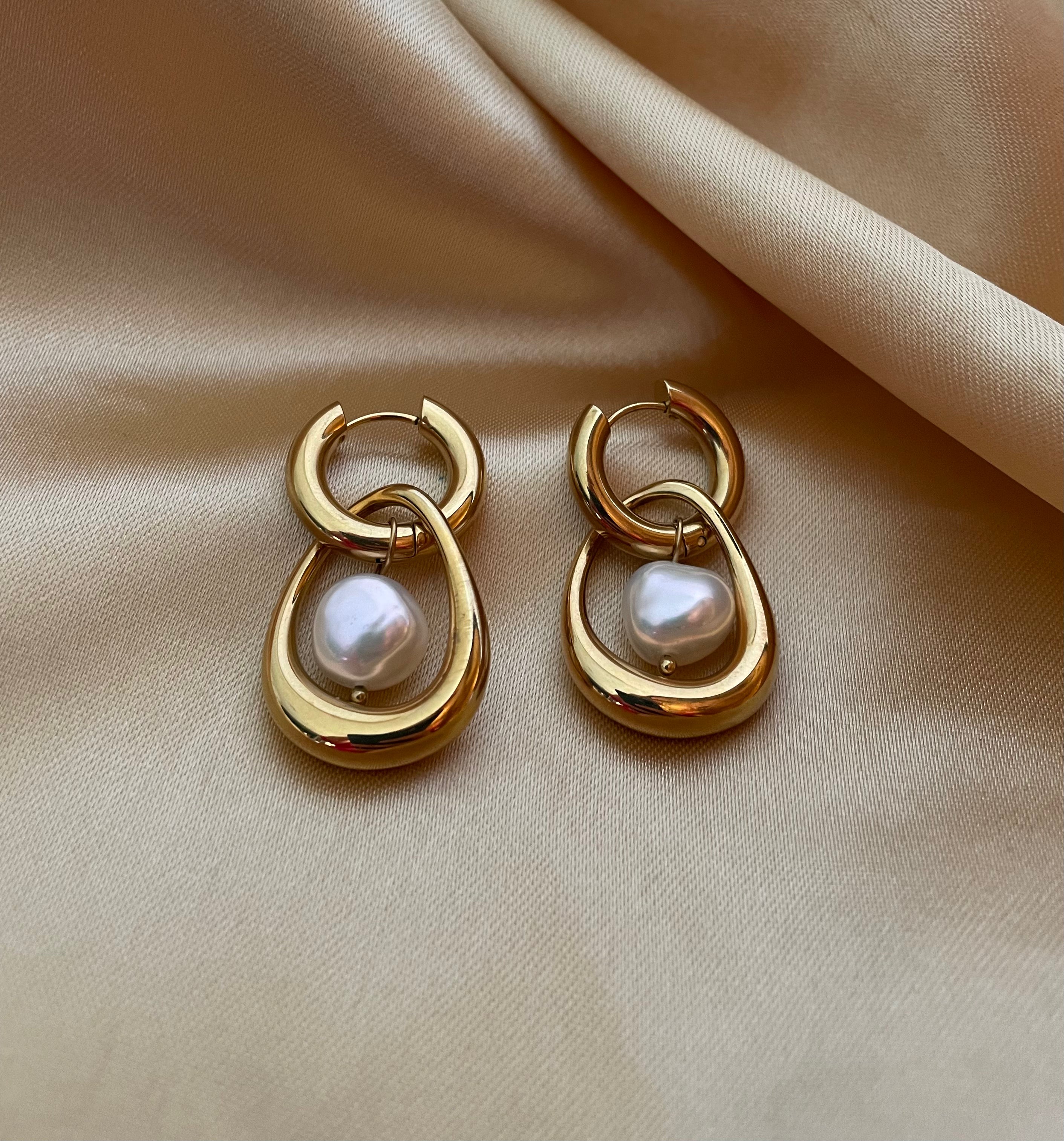 Close-up editorial photography of a gold hoop drop earring featuring a suspended white high-luster pearl, worn by a model with blonde-brown hair against a matte ivory background. High-resolution luxury jewelry shot highlighting realistic gold texture and pearl iridescence.
