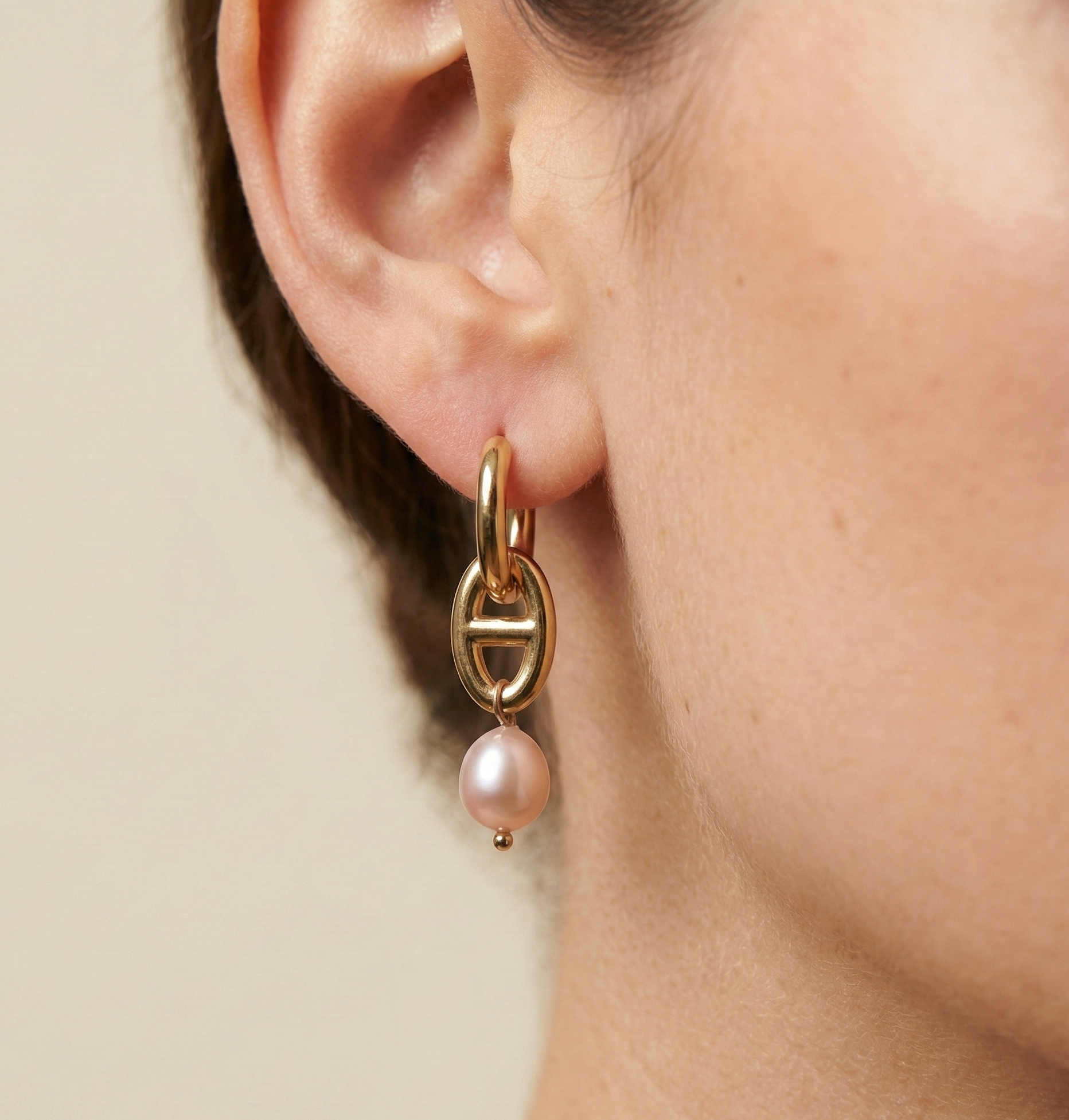 Gold hoop earring with a pink pearl on an ear against a neutral background