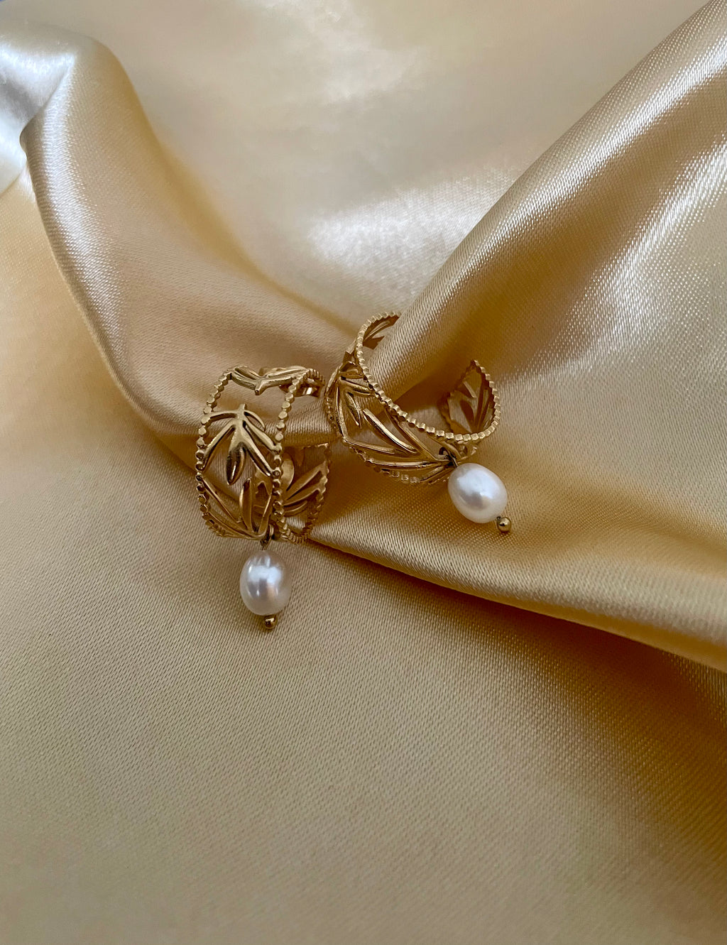 Close-up of a detailed gold openwork hoop earrings featuring a leaf motif and a single dangling perfect white oval pearl. The warm, soft lighting highlights the pearl's luster and the gold texture, set against a neutral background.