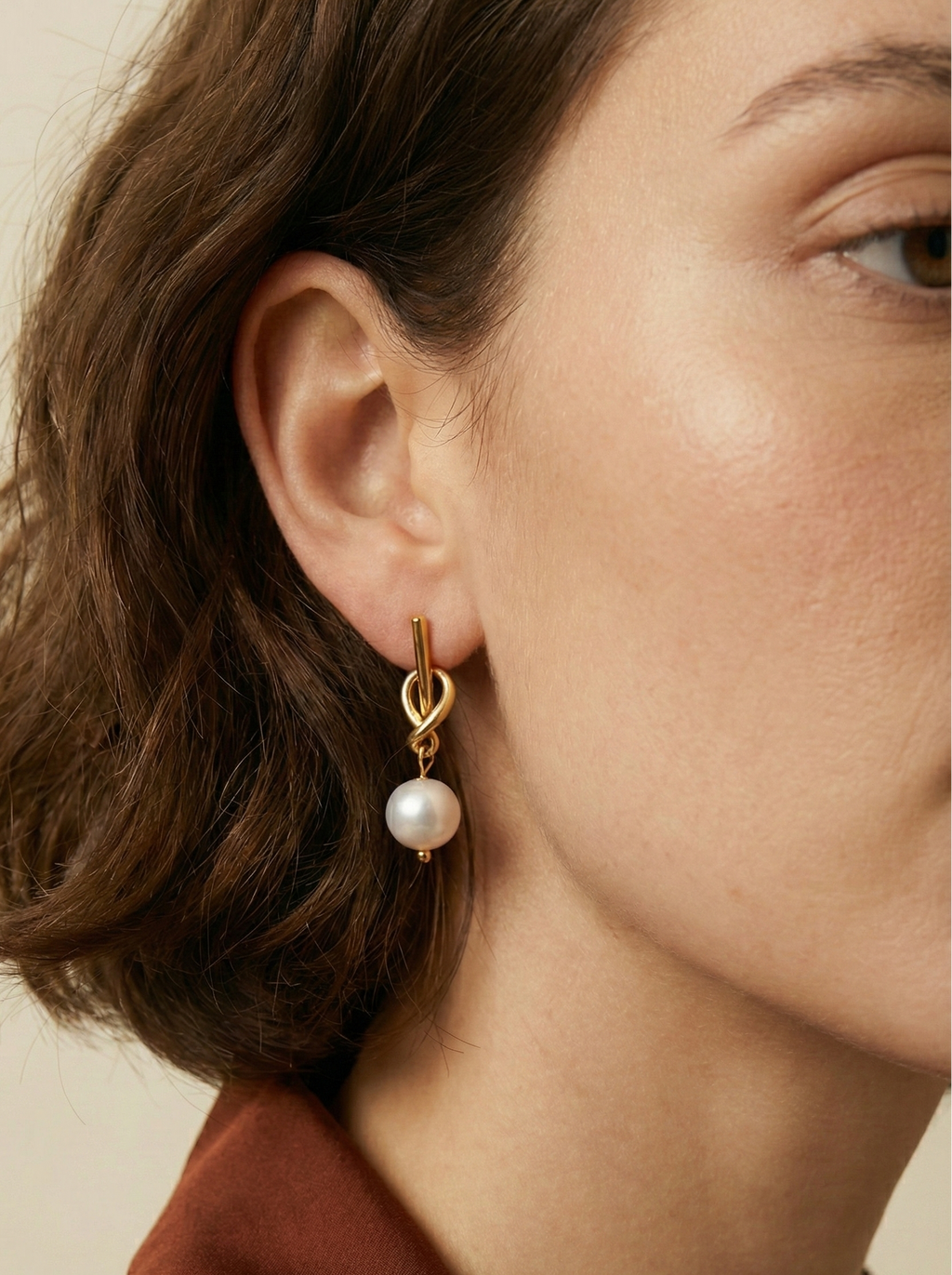 front profile of a woman with short wavy hair wearing elegant gold knot earrings with a suspended white pearl. The lighting highlights the luster of the pearl and the gold finish