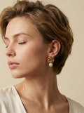 Close-up of gold twisted knot earrings with a white pearl drop, worn by a model in a cream silk blouse