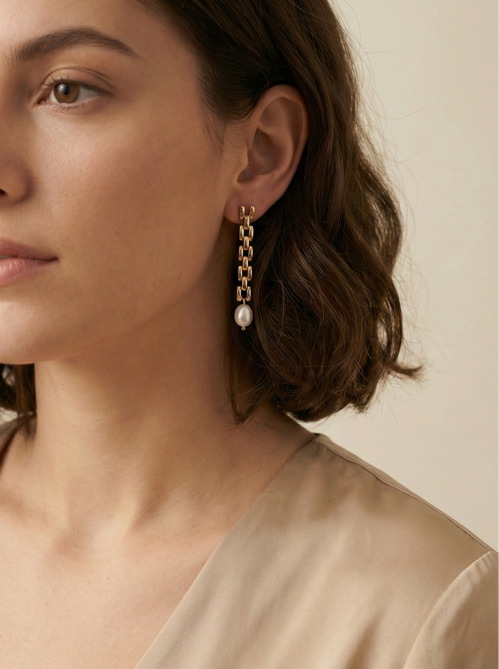 Close-up of a woman wearing gold chain earrings with pearls against a neutral background