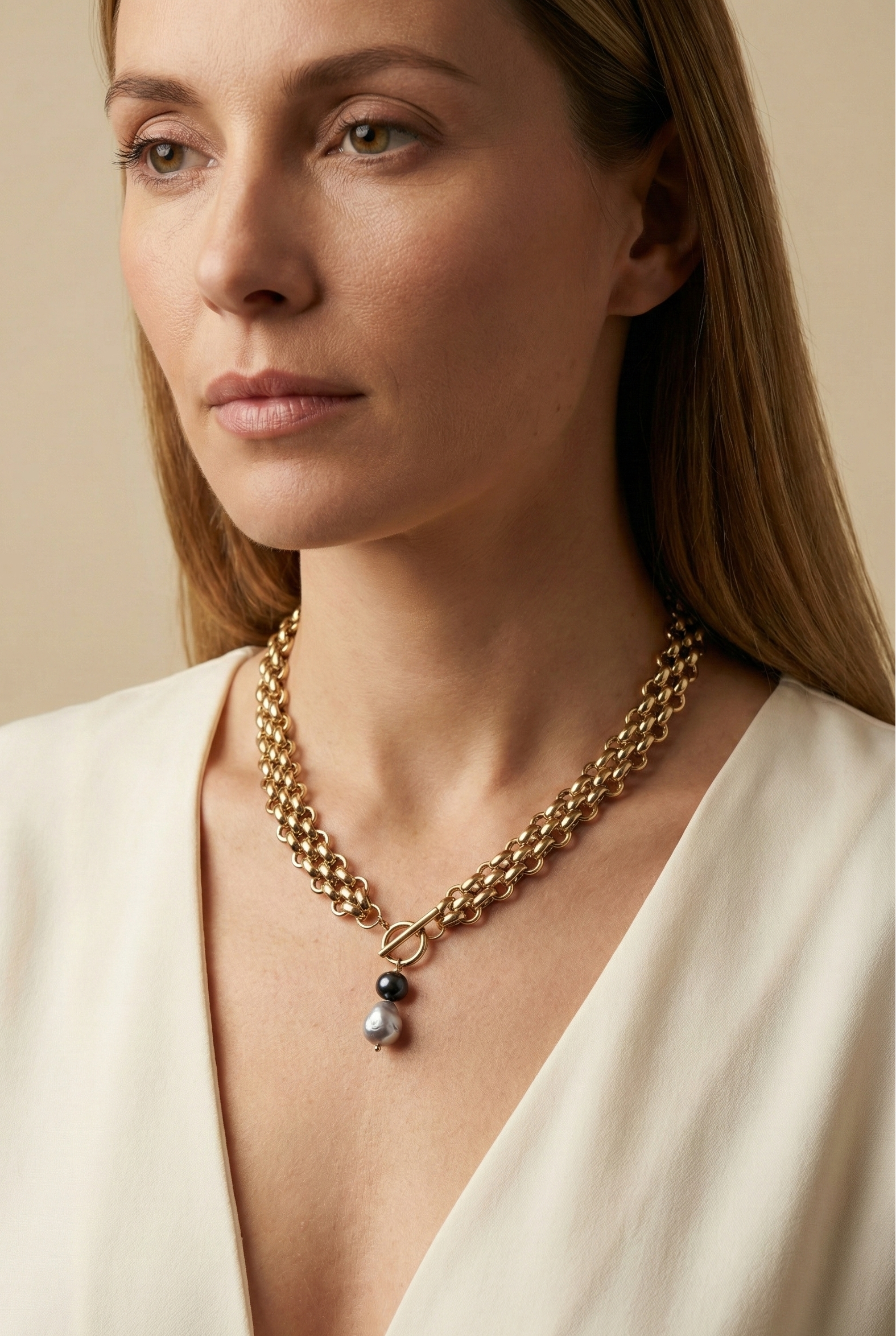 Elegant French model tucking hair behind ear wearing a chunky gold link chain necklace with a T-bar toggle clasp, featuring a drop pendant with a round black pearl and a grey baroque pearl against an ivory top