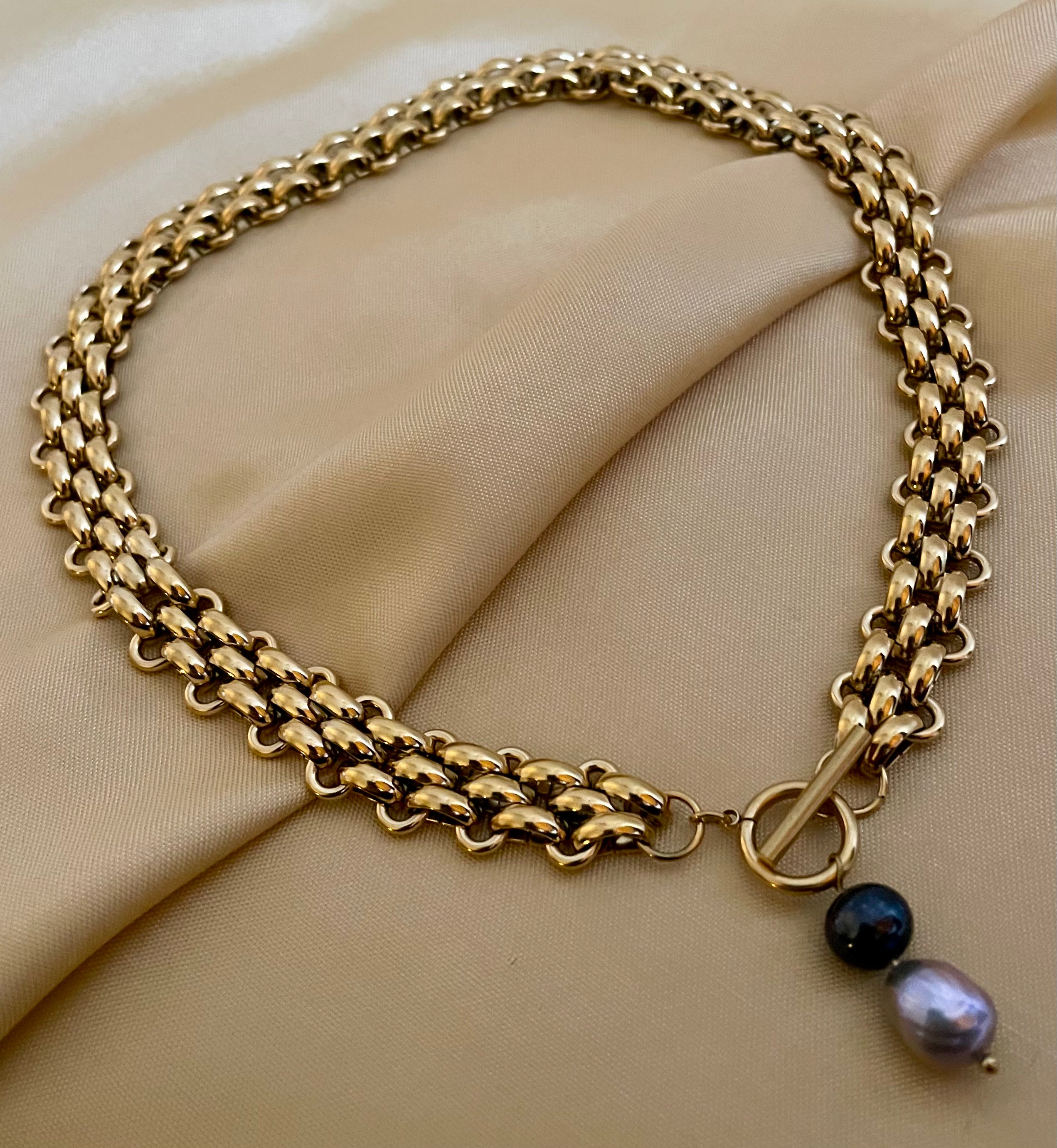 Elegant French model tucking hair behind ear wearing a chunky gold link chain necklace with a T-bar toggle clasp, featuring a drop pendant with a round black pearl and a grey baroque pearl against an ivory top