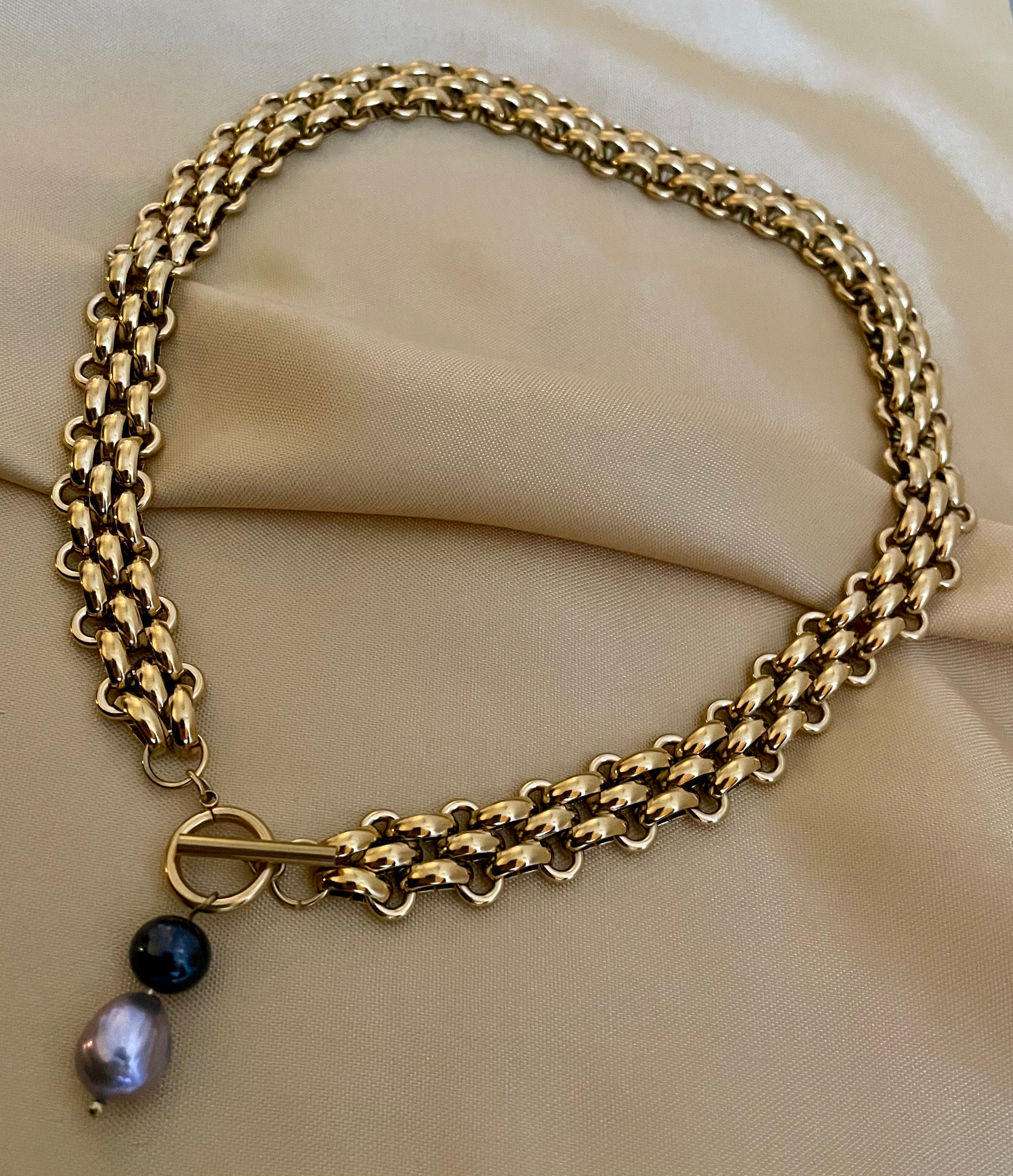 chunky gold link chain necklace with a T-bar toggle clasp, featuring a drop pendant with a round black pearl and a grey baroque pearl against an ivory top