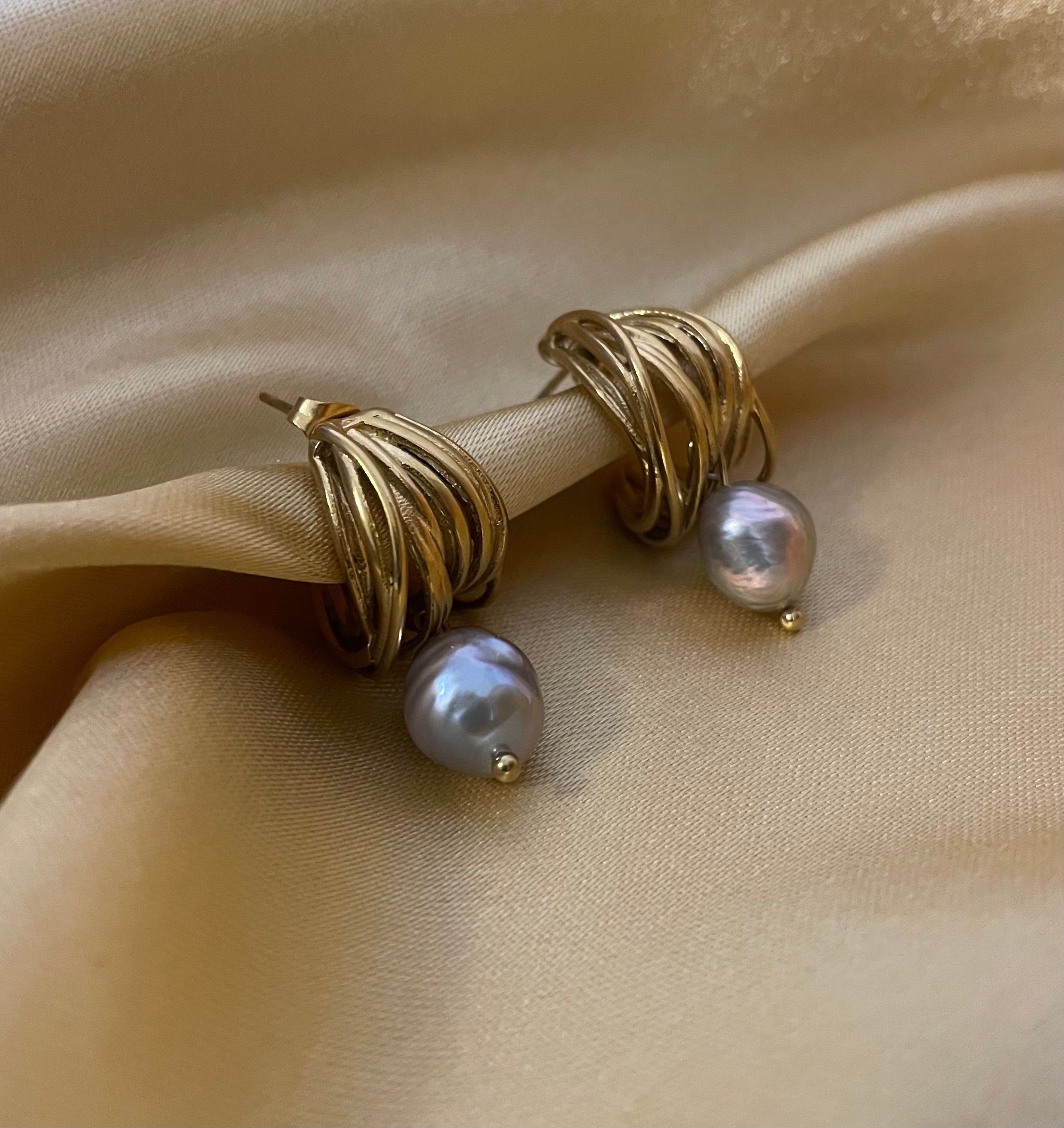 Close-up of 18k gold multi-hoop earrings featuring a lustrous grey pearl drop. The jewelry highlights a textured gold finish and organic pearl shape, set against a plain ivory background