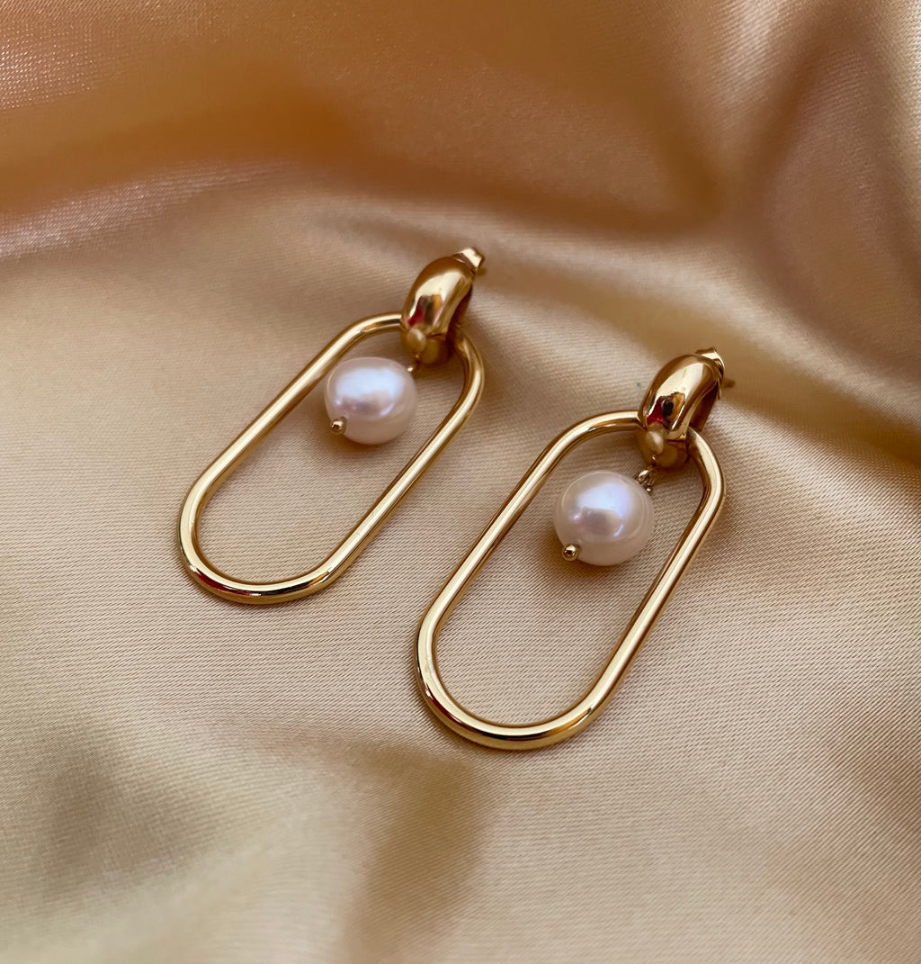 Close-up view of gold elongated oval hoop earrings featuring a suspended white baroque pearl, shown worn on the ear to verify size and scale