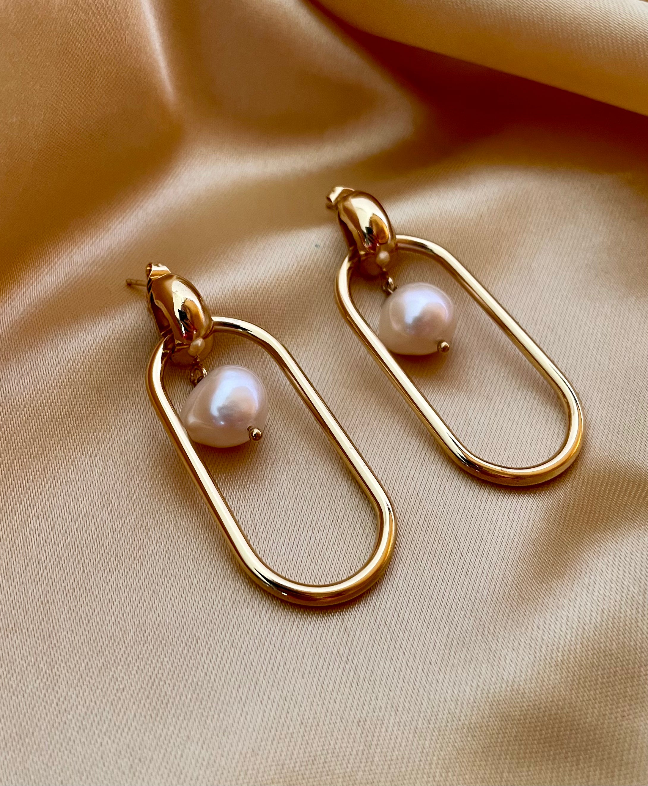 Close-up view of gold elongated oval hoop earrings featuring a suspended white baroque pearl, shown worn on the ear to verify size and scale