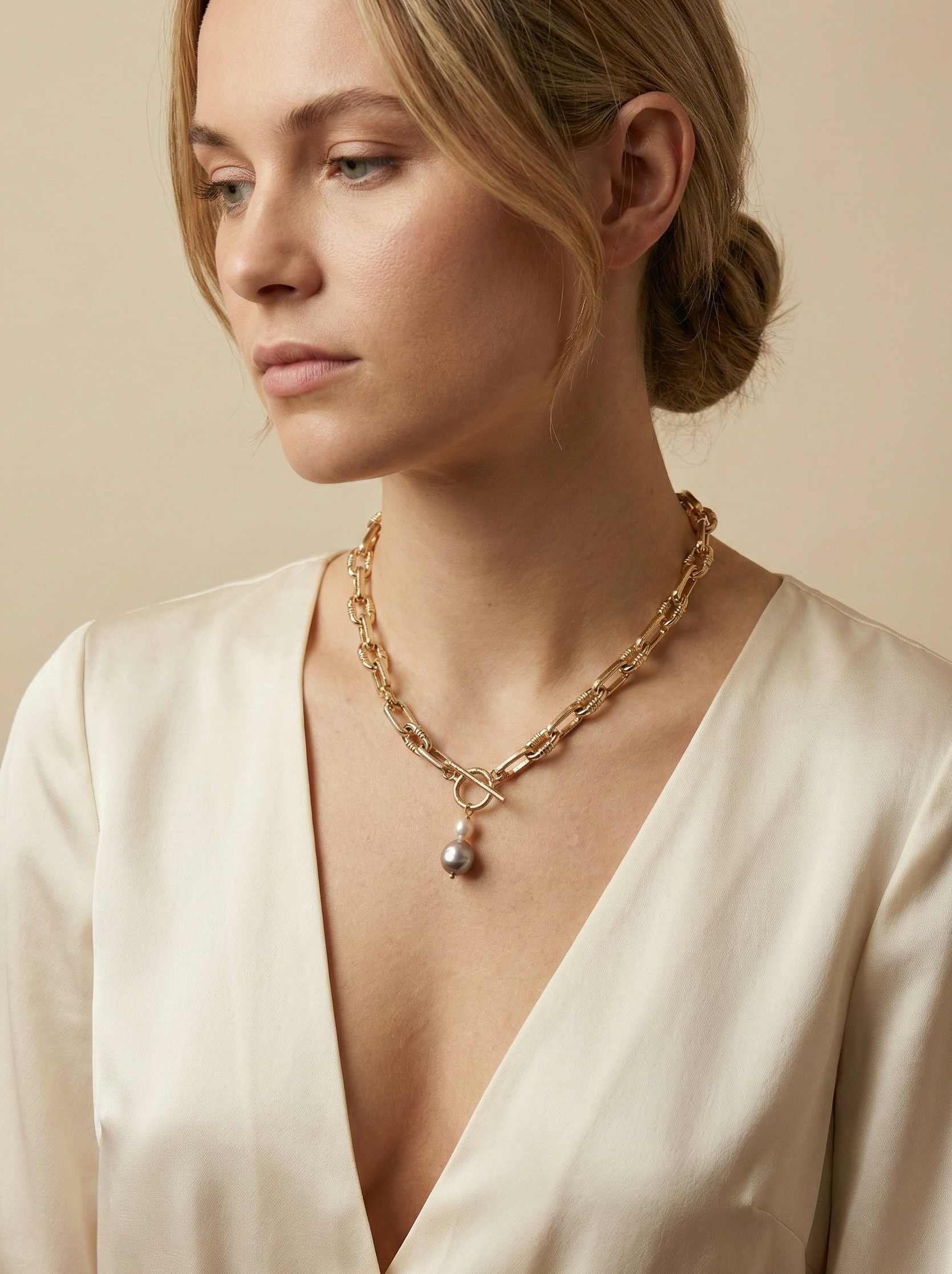 Detail shot of a luxury gold link necklace worn on an ivory silk top. The focal point is a T-bar toggle closure holding a high-shine metallic grey baroque pearl and a champagne oval pearl, showing deep luster and smooth nacre